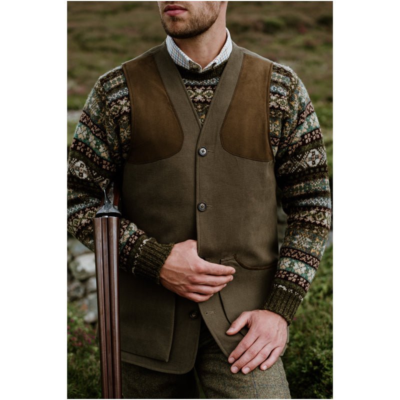 A man wears a Campbells of Beauly Cotton Shooting Waistcoat with a patterned knit sweater and tweed trousers, standing outdoors holding a shotgun—perfectly attired for country pursuits. His face is partially out of frame.