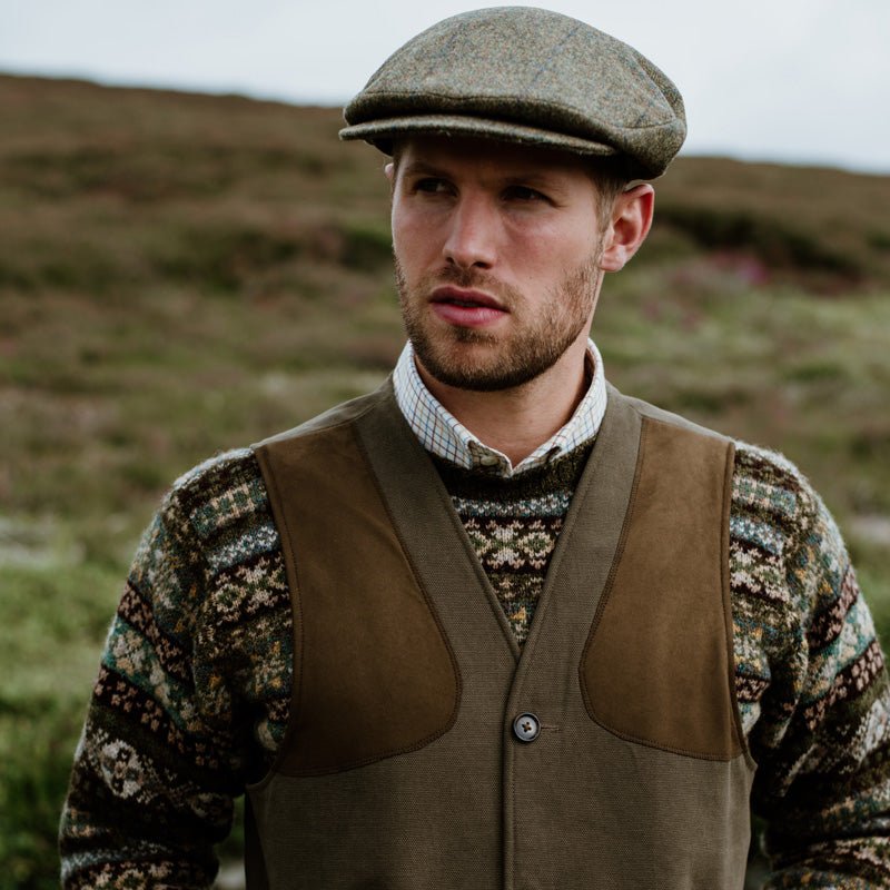 A man wearing a patterned sweater, a brown shooting vest, a checked shirt, and a tweed flat cap stands outdoors in a grassy, hilly landscape, looking slightly to the side.
