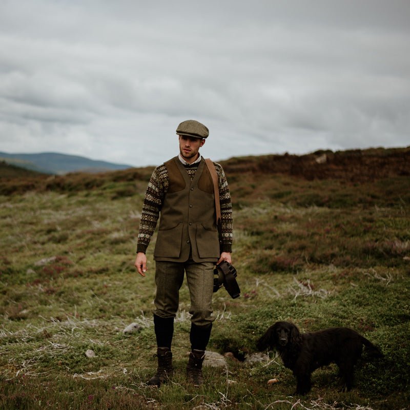 A man in country attire and flat cap walks the grassy moorland with a black dog, wearing a Campbells of Beauly Cotton Shooting Waistcoat over a patterned sweater as he enjoys countryside pursuits beneath cloudy skies.