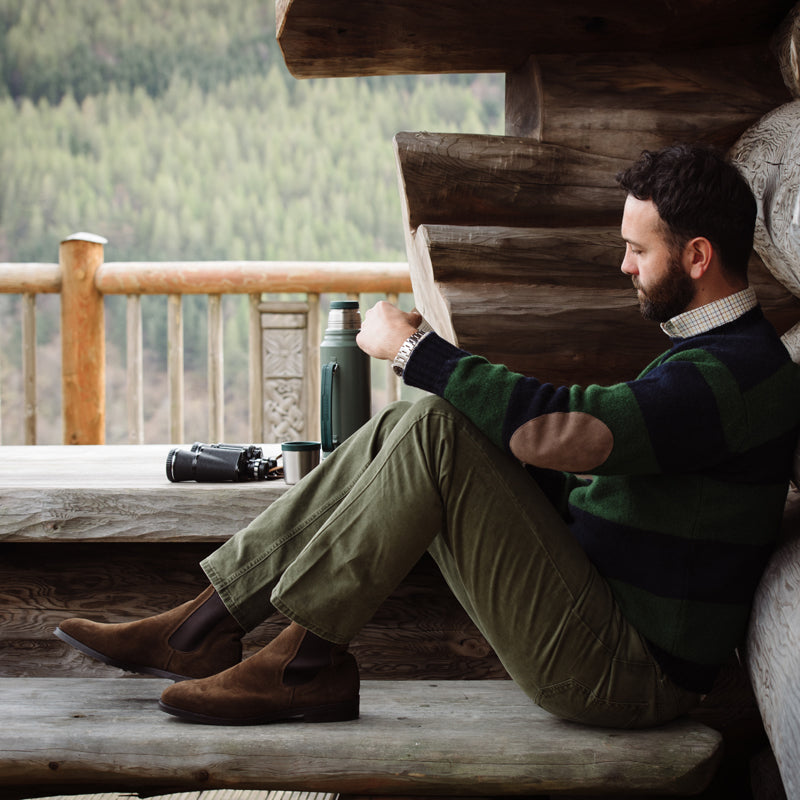 A bearded man on a wooden bench by a log cabin wears a striped sweater, olive pants, and Campbell’s of Beauly Suede Chelsea Boots. He checks his watch; binoculars and a green thermos are nearby, with forest views behind him.