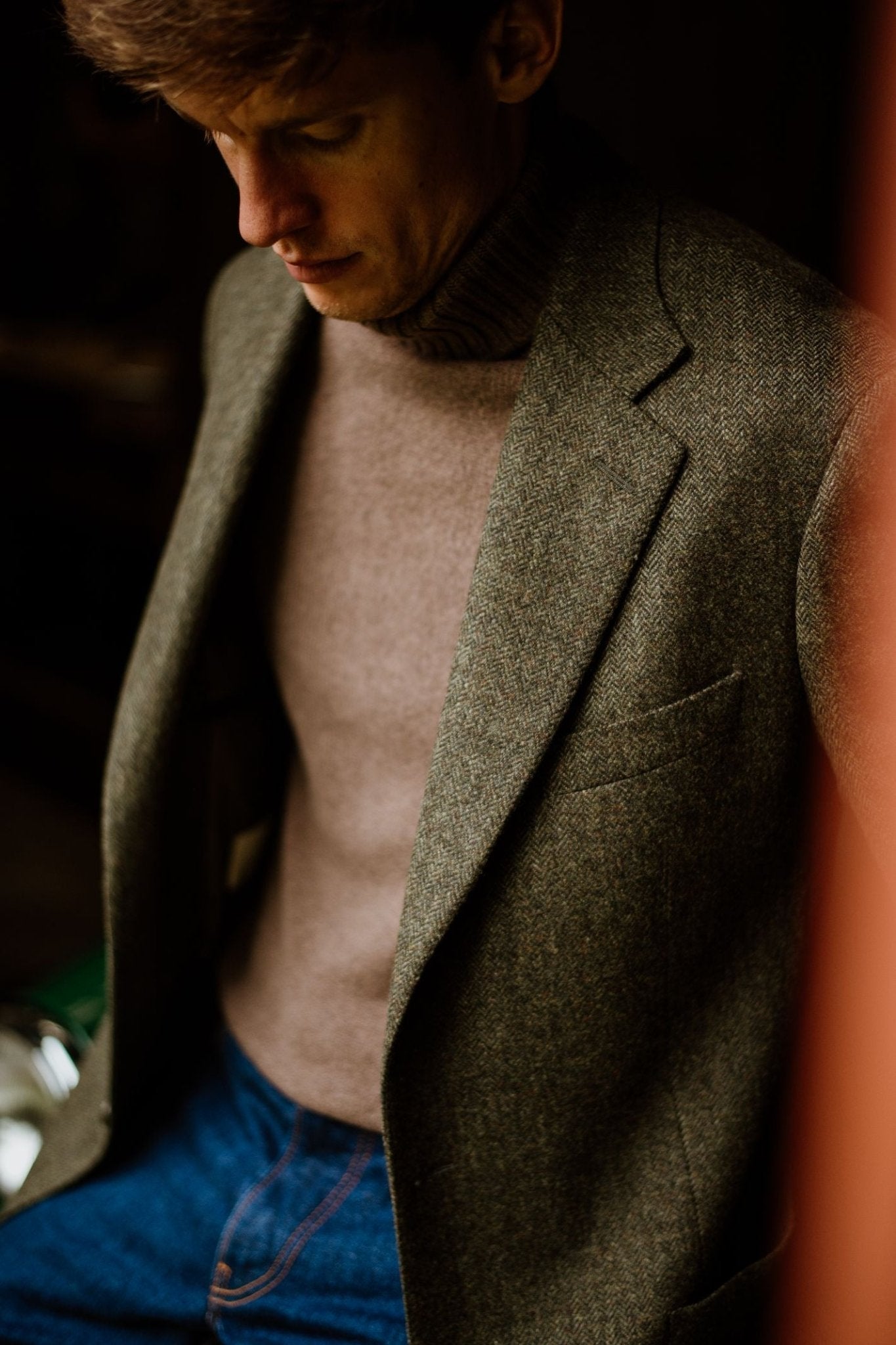 A man gazes down, wearing a Campbells of Beauly Forest Herringbone House Jacket over a brown turtleneck and blue jeans, softly illuminated.