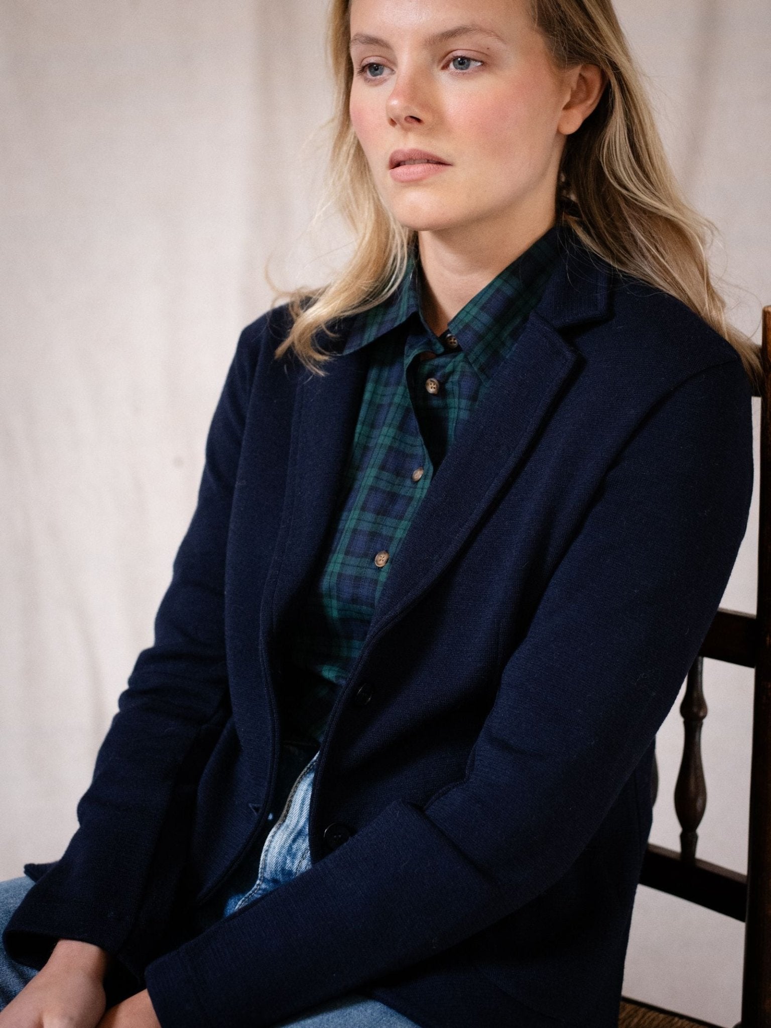 A woman with long blonde hair sits on a wooden chair, wearing Campbell's of Beauly Merino Blazer in navy over a green and blue plaid shirt and light blue jeans, set against a plain light background.