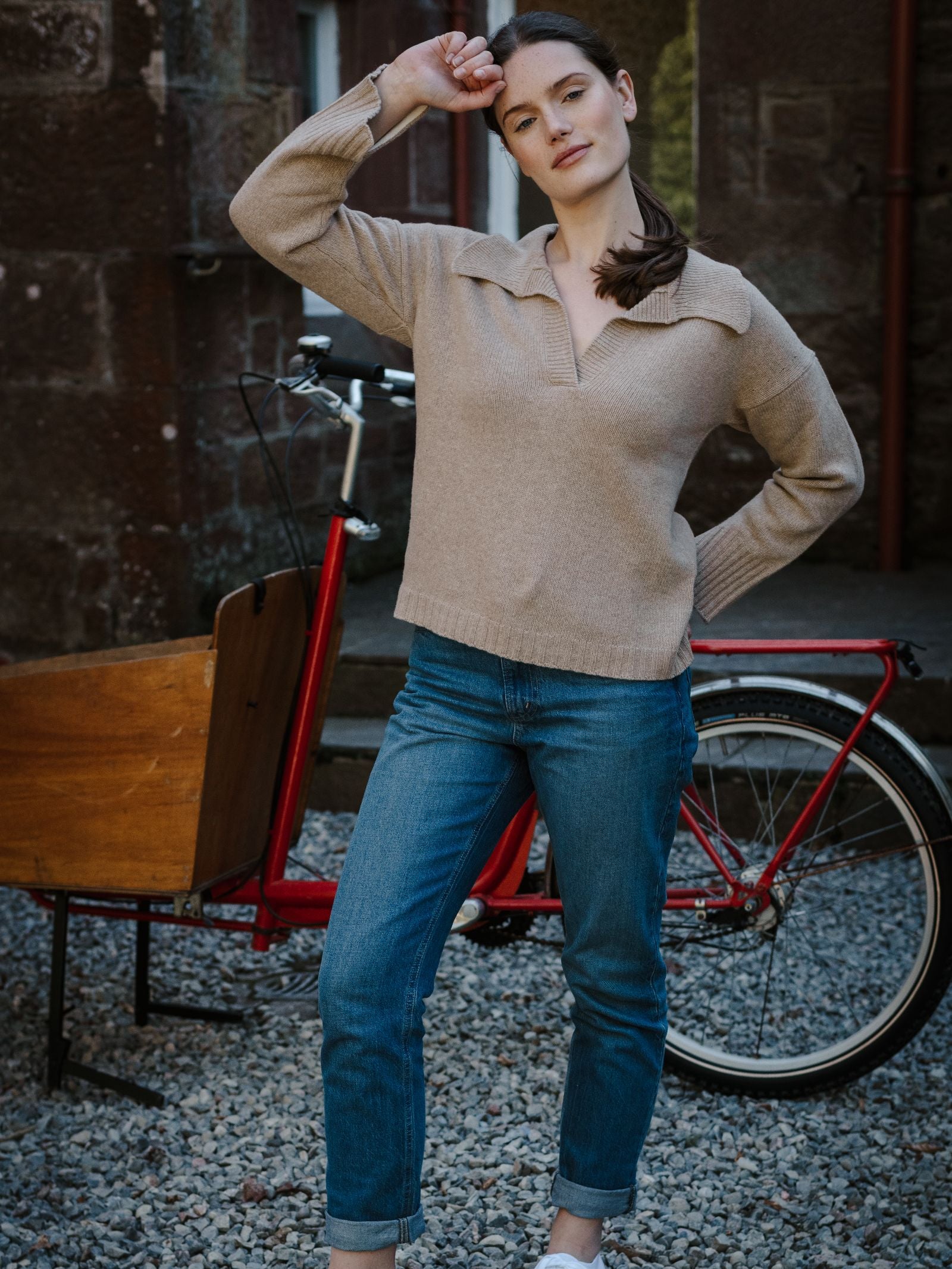 A woman wears the Campbell’s of Beauly Merino Cashmere Shirt Neck Jumper in beige with blue jeans, standing before a red bicycle with a wooden cargo box, one arm raised, against a stone wall and gravel ground.
