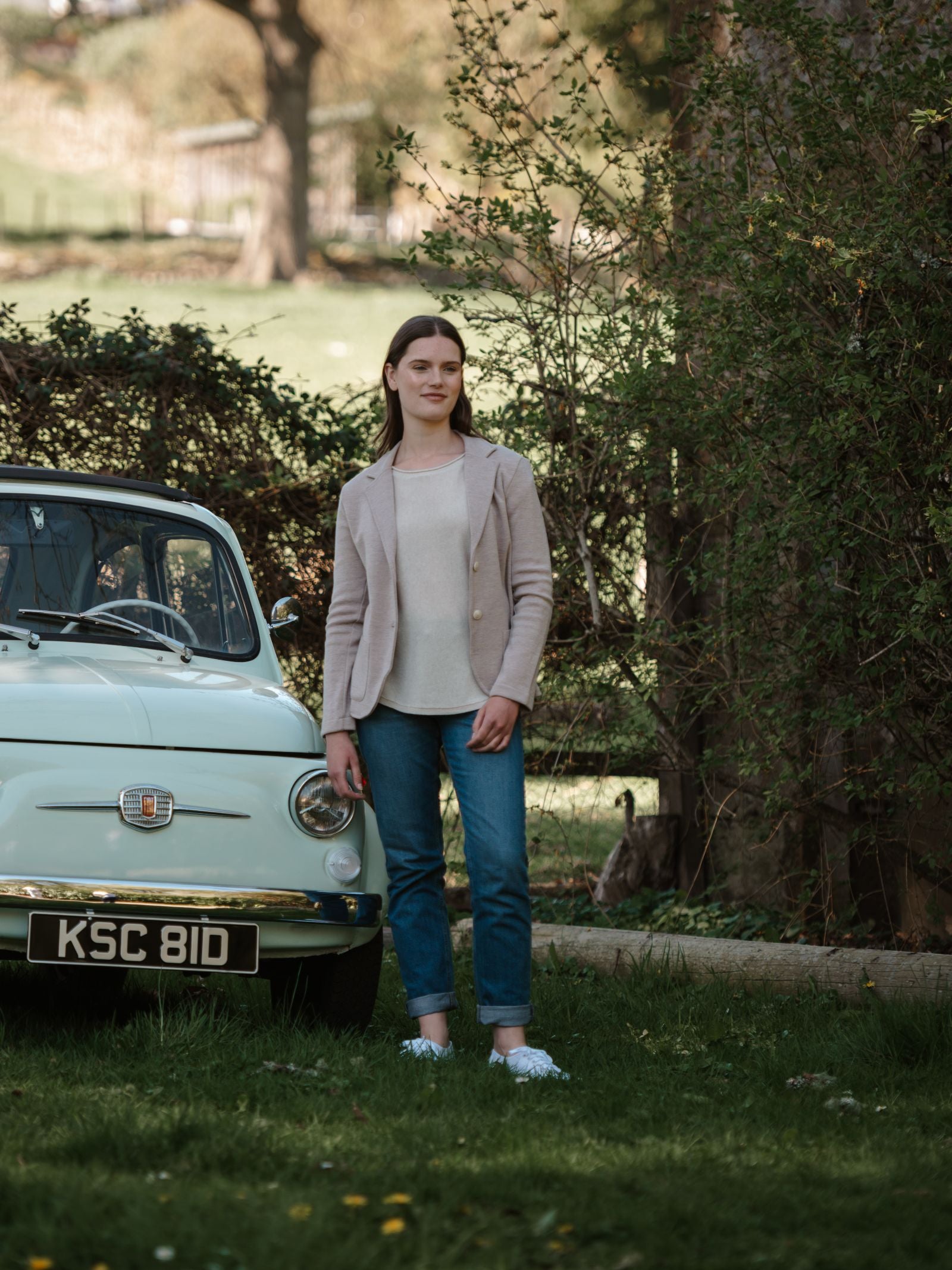 A woman in a Campbell's of Beauly Merino Blazer stands on grass by a vintage light blue Fiat KSC 81D. She pairs the made-in-Italy jacket with a white top, blue jeans, and white shoes. Trees and bushes fill the background.