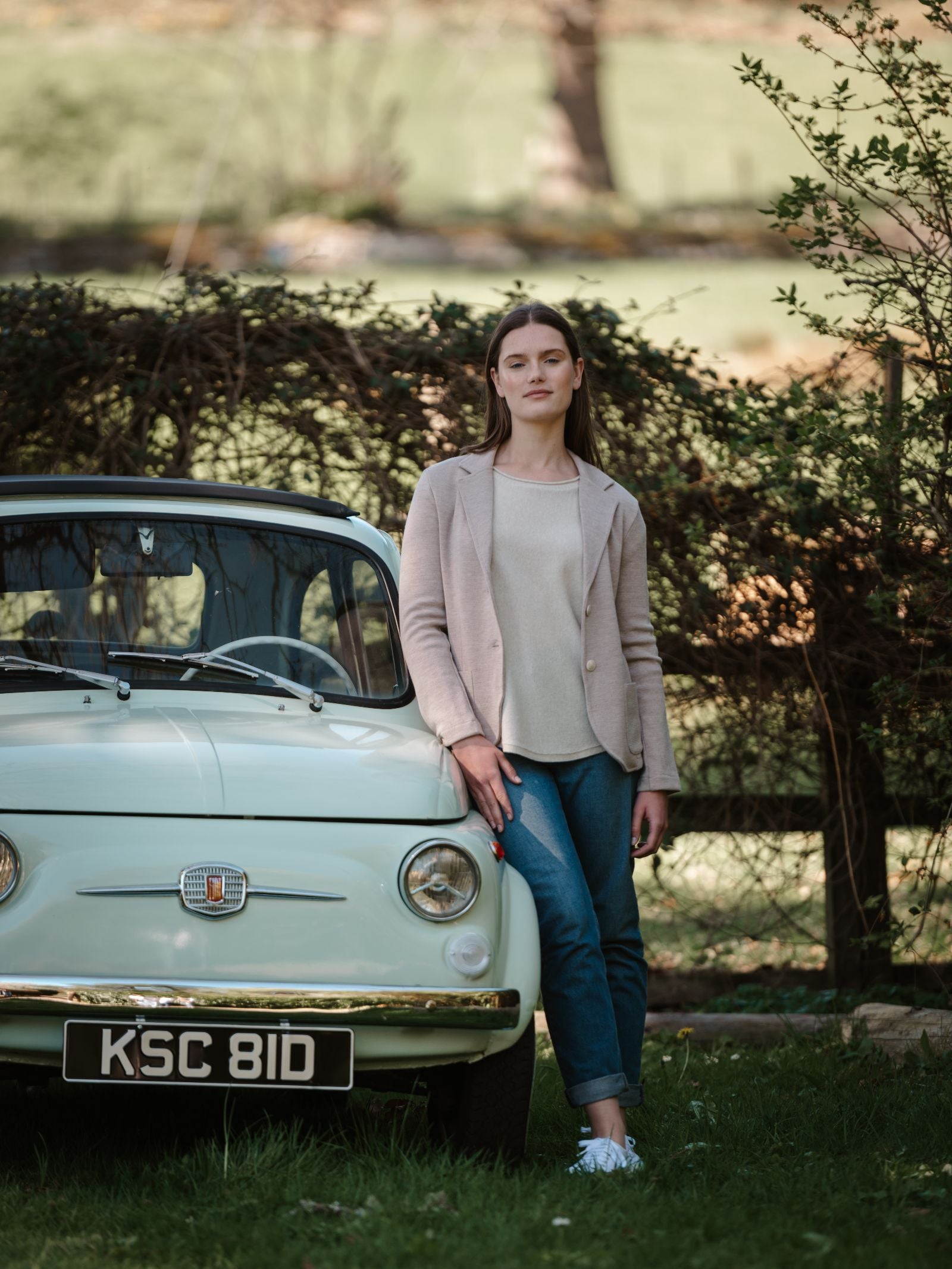 A woman in the Campbell's of Beauly Merino Blazer, a beige top, and blue jeans stands beside a vintage pale green Fiat outdoors, surrounded by greenery and a wooden fence.