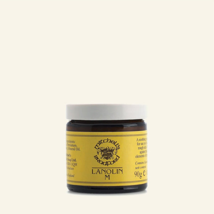 A brown glass jar with a yellow label reading “Lanolin Salve” by Mitchell’s of Bradford. It has a sheep logo, white screw-on lid, 90g content, and is ideal for sensitive skin. The jar sits on a plain, light cream background.