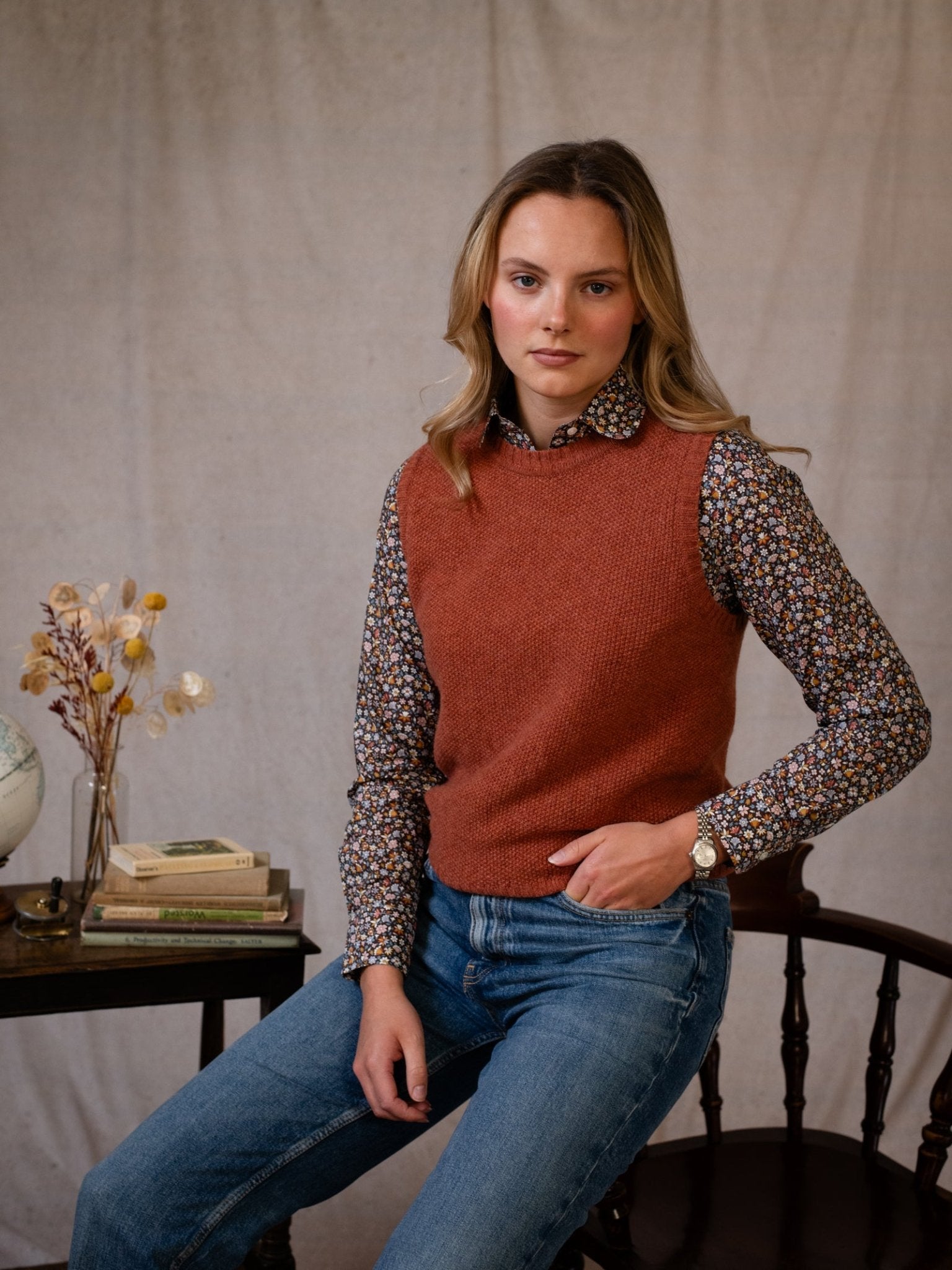 A woman with long blonde hair sits on a wooden chair, wearing the Campbell's of Beauly Moss Stitch Crew Tank over a floral shirt and blue jeans. She rests her hand on her knee; books and a vase are visible on the table behind her.