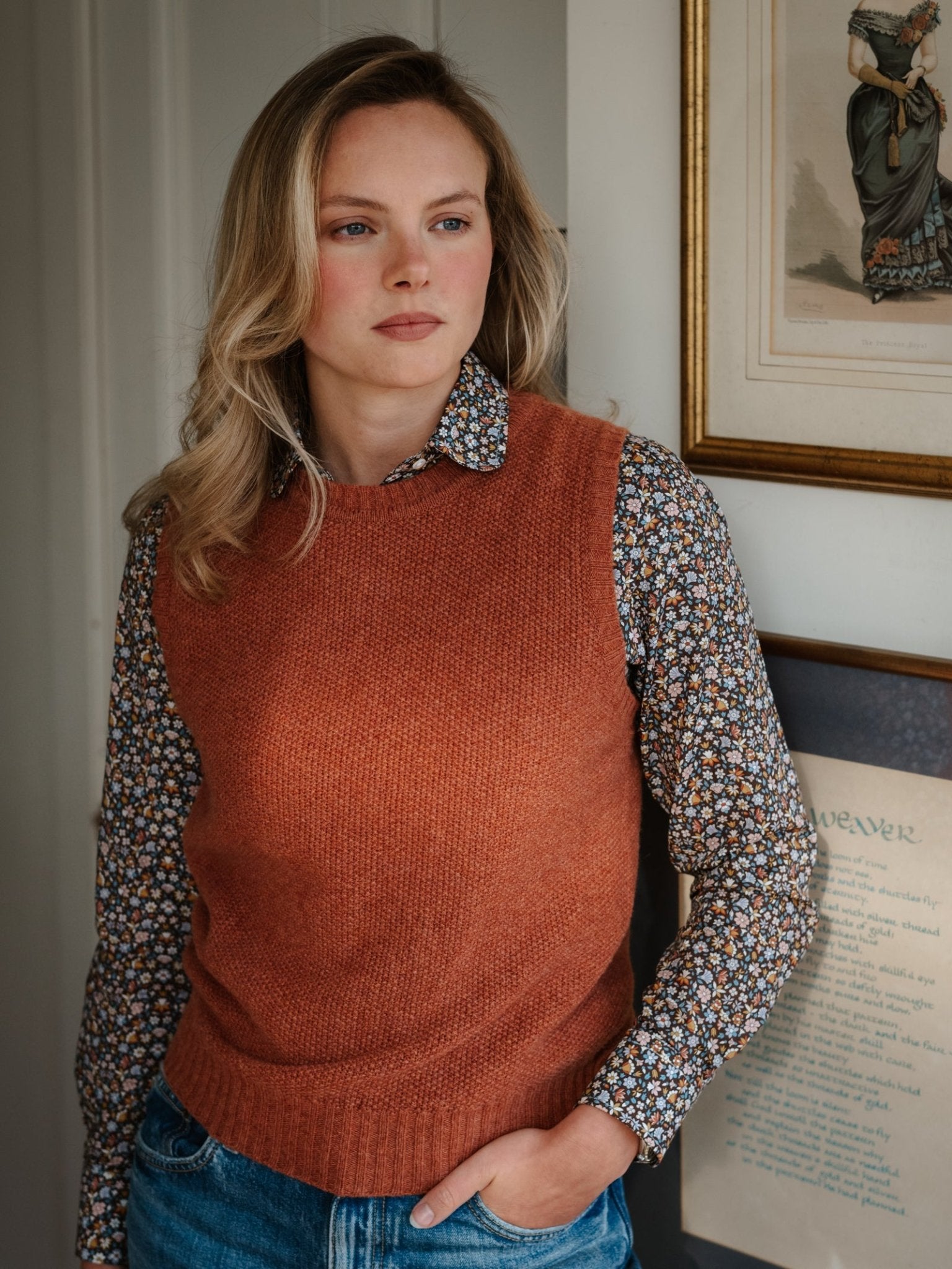 A woman with wavy blonde hair and jeans stands indoors near framed pictures, wearing a floral shirt under the Campbell's of Beauly Moss Stitch Crew Tank, with one hand in her pocket and a thoughtful expression.