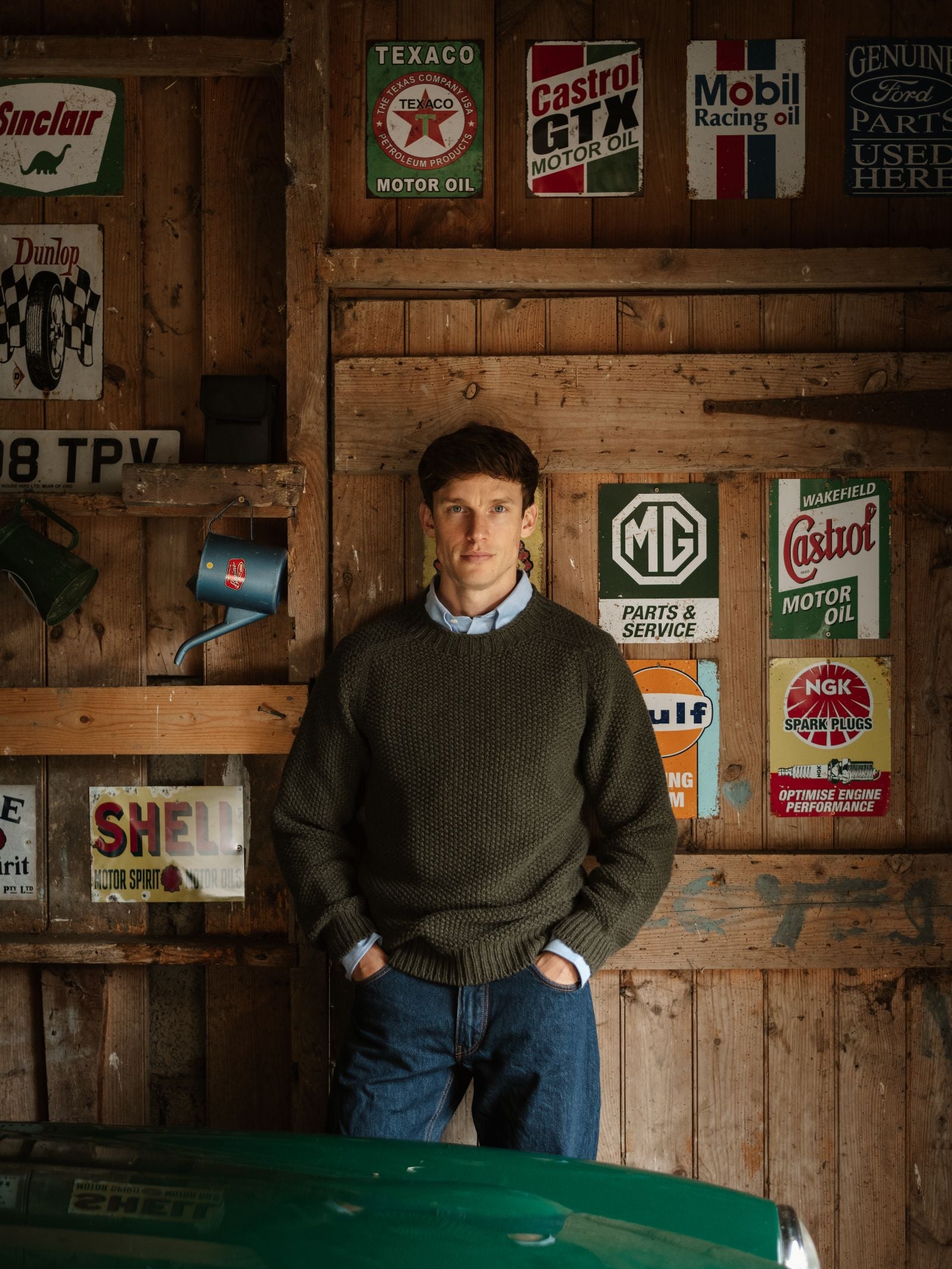A man wearing a classic Moss Stitch Jumper by Campbell's of Beauly and jeans stands in a wooden garage with vintage motor oil and car signs behind him, while part of a green car is visible in the foreground.