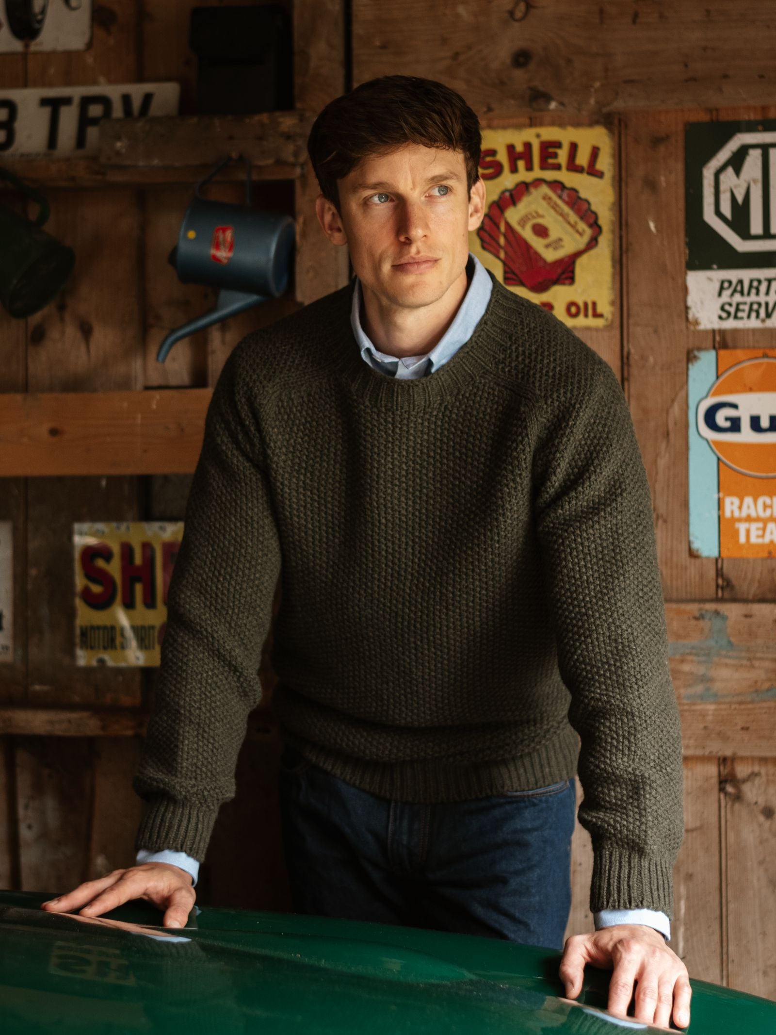A man with short brown hair wears a Campbell's of Beauly Moss Stitch Jumper and a blue shirt, standing with hands on a green surface in a rustic garage decorated with vintage signs on wooden walls.