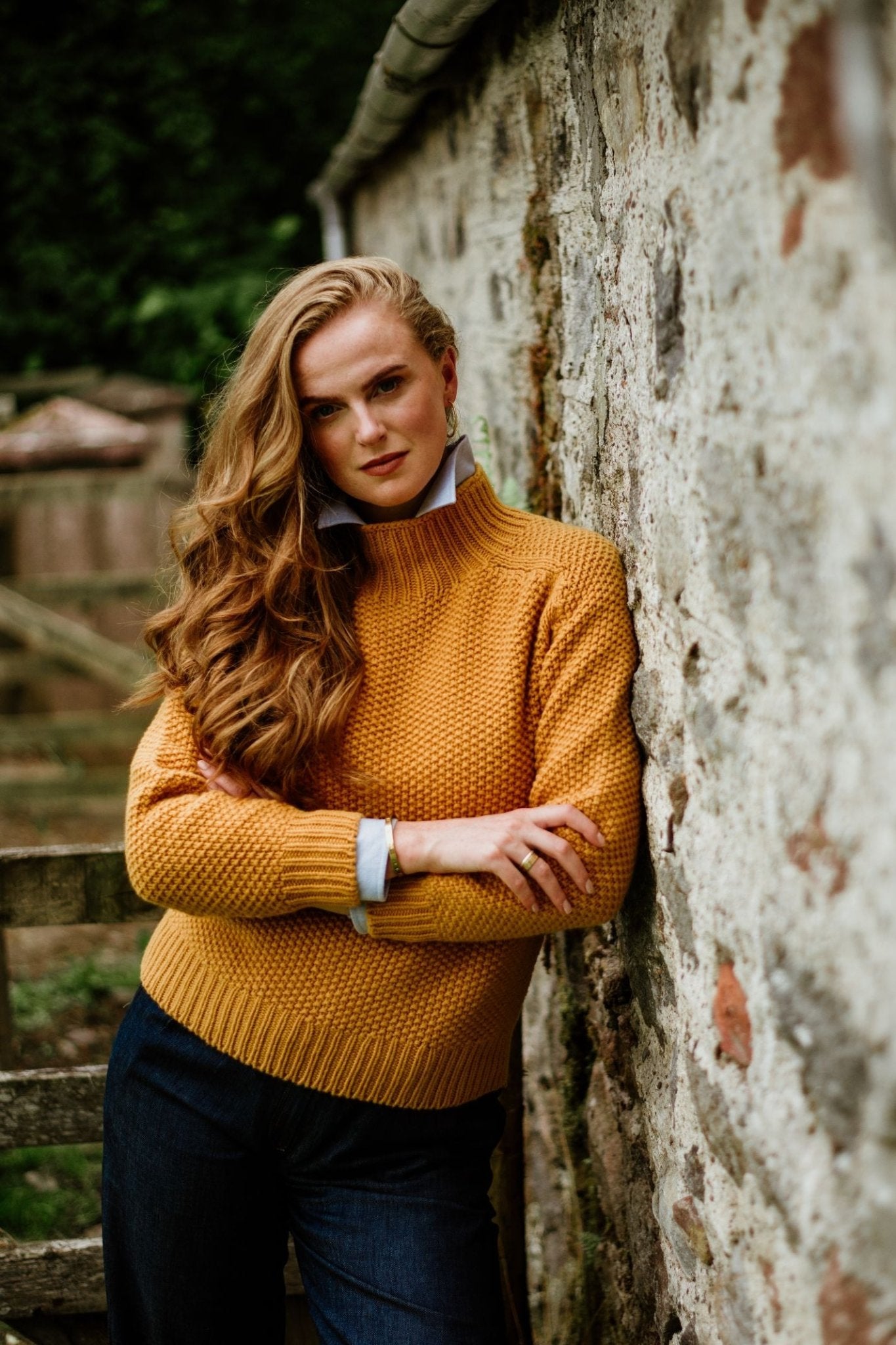 A woman with long, wavy red hair in a Campbells of Beauly Chunky Moss Stitch Polo and dark jeans leans against a rustic stone wall outdoors, arms crossed, looking confidently at the camera.