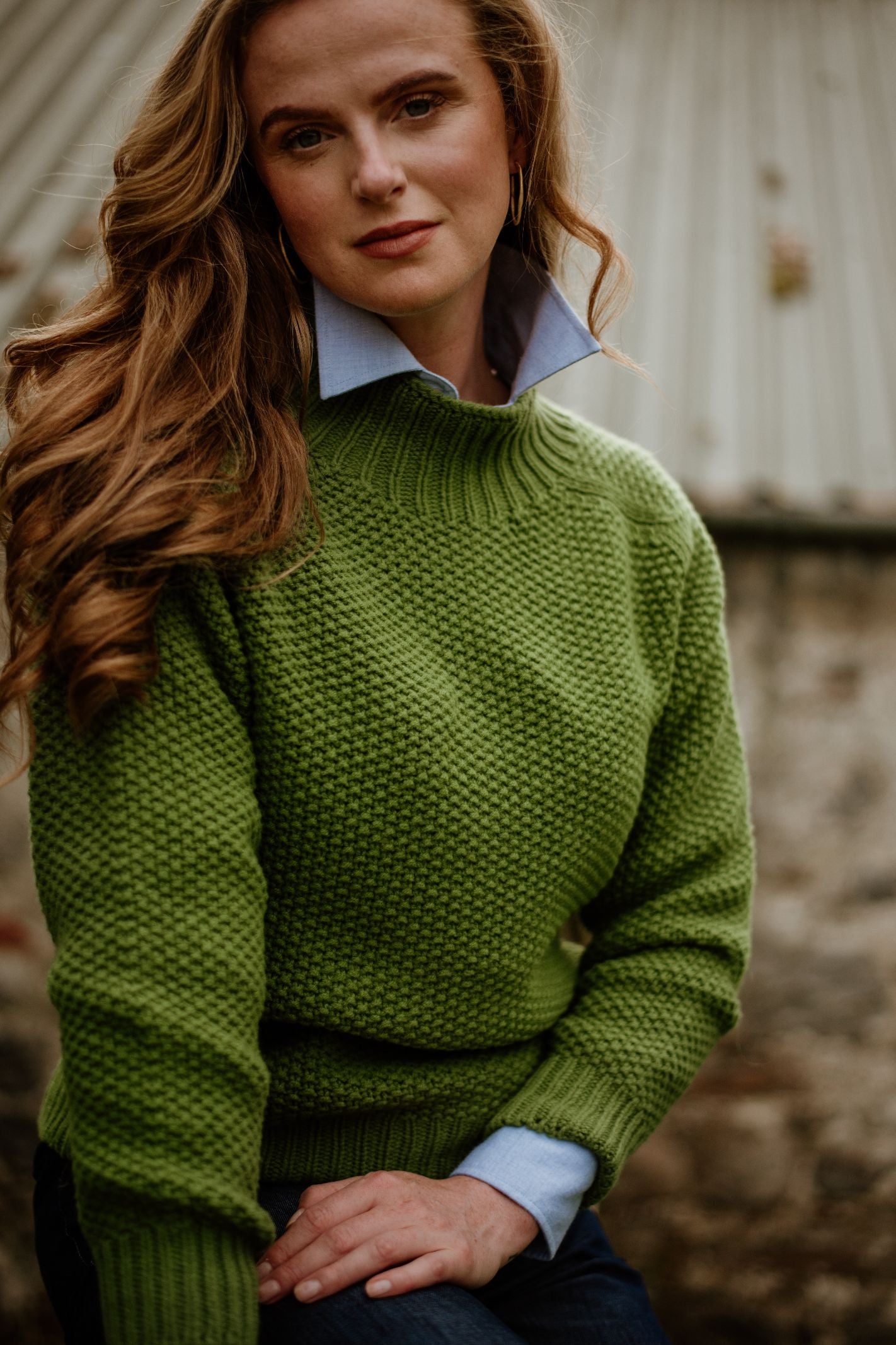 A woman with long wavy hair wears the Campbells of Beauly Chunky Moss Stitch Polo sweater in green over a blue collared shirt, sitting outdoors before a rustic corrugated metal-roofed building, gazing calmly at the camera.
