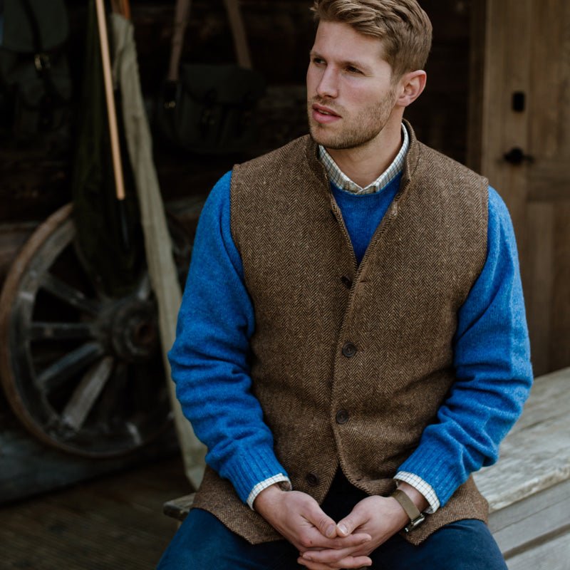 A man in a blue sweater, checked shirt, and Campbell’s of Beauly Tweed Nehru Gilet sits on a wooden bench outdoors by a wagon wheel and rustic wall, gazing thoughtfully into the distance.