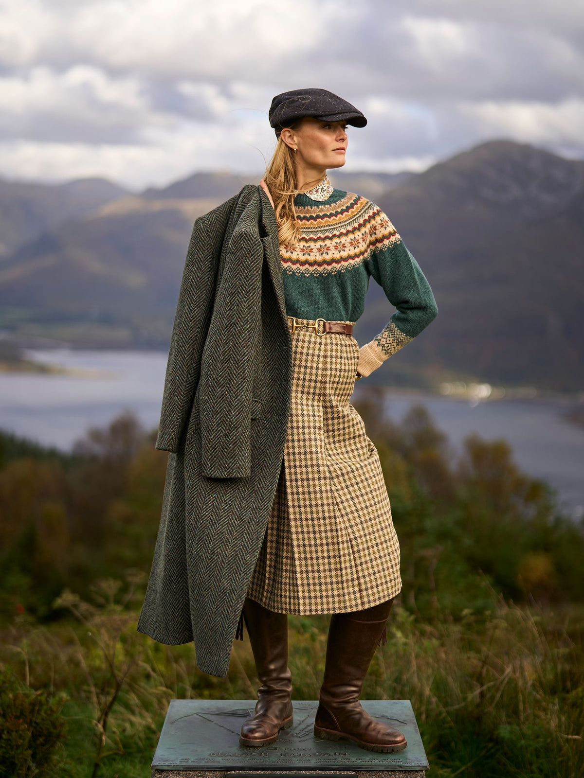 A woman wearing a patterned green sweater, a tan and green Dogtooth Skirt from Campbell’s of Beauly, and brown leather boots stands outdoors holding a long coat over her shoulder.
