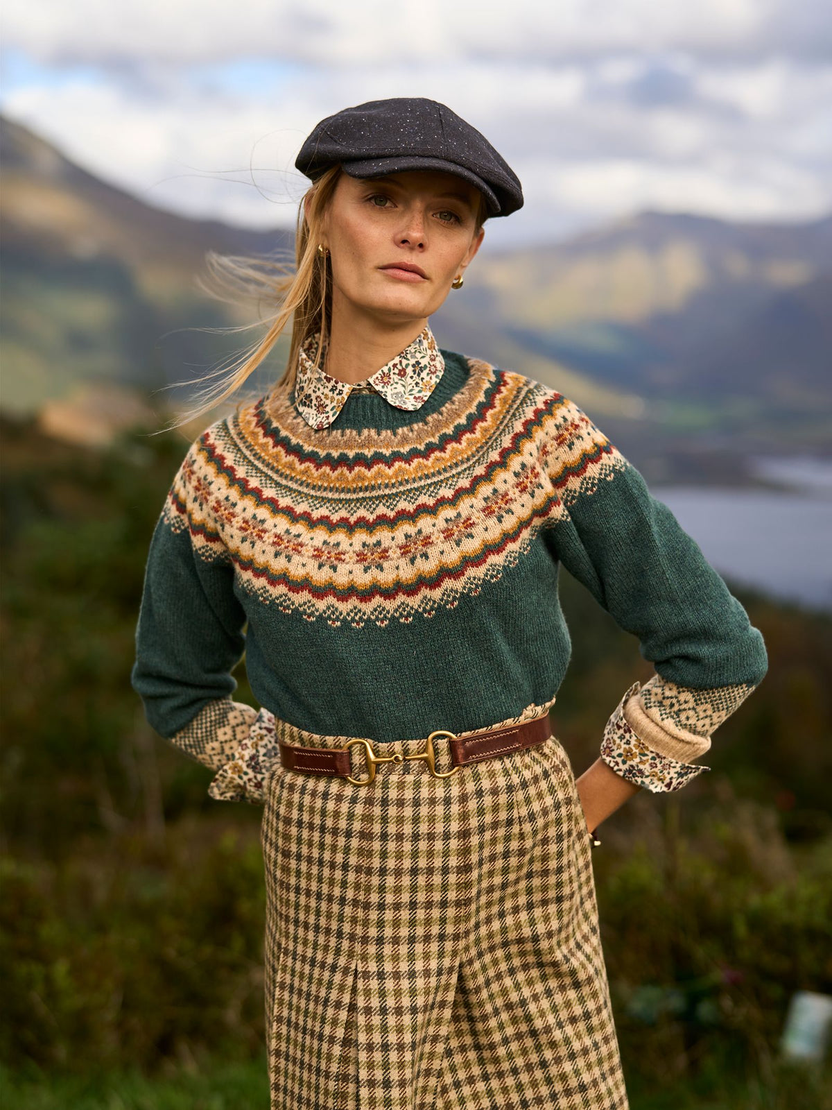 A woman wearing a dark green Fair Isle sweater over a floral shirt and a Campbell’s of Beauly Dogtooth Skirt, accessorized with a brown leather belt and a flat cap.