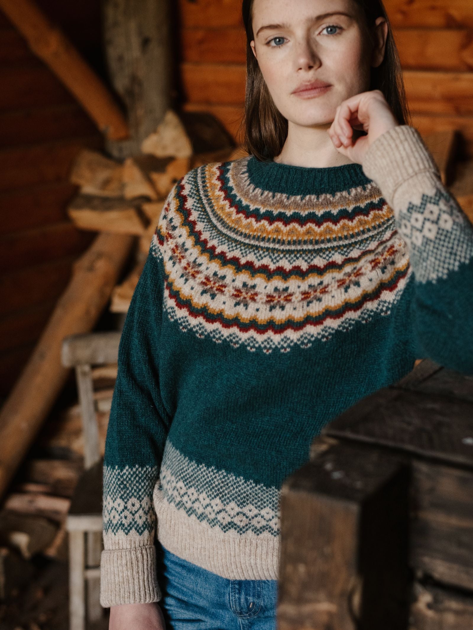 A woman wearing the Campbell's of Beauly Nordic Fairisle Crew Jumper stands in a rustic wooden room with firewood and wooden furniture, looking at the camera with a neutral expression.