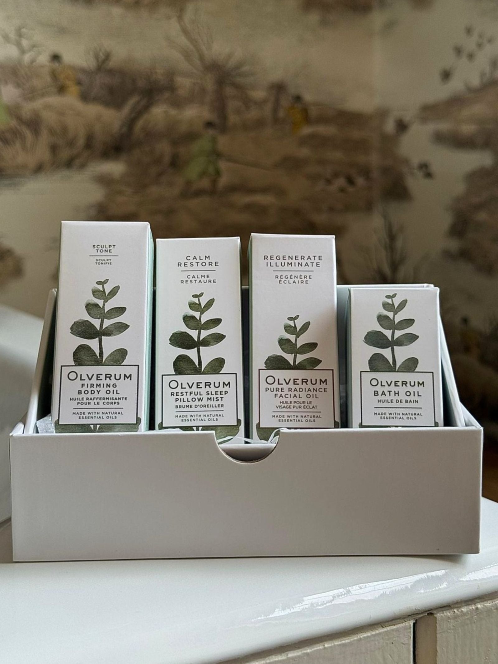 A white display box, the OLVERUM Discovery Box, holds four skincare products with botanical illustrations and text, including bath oil and firming body oil, set against nature-themed wallpaper on a white surface.