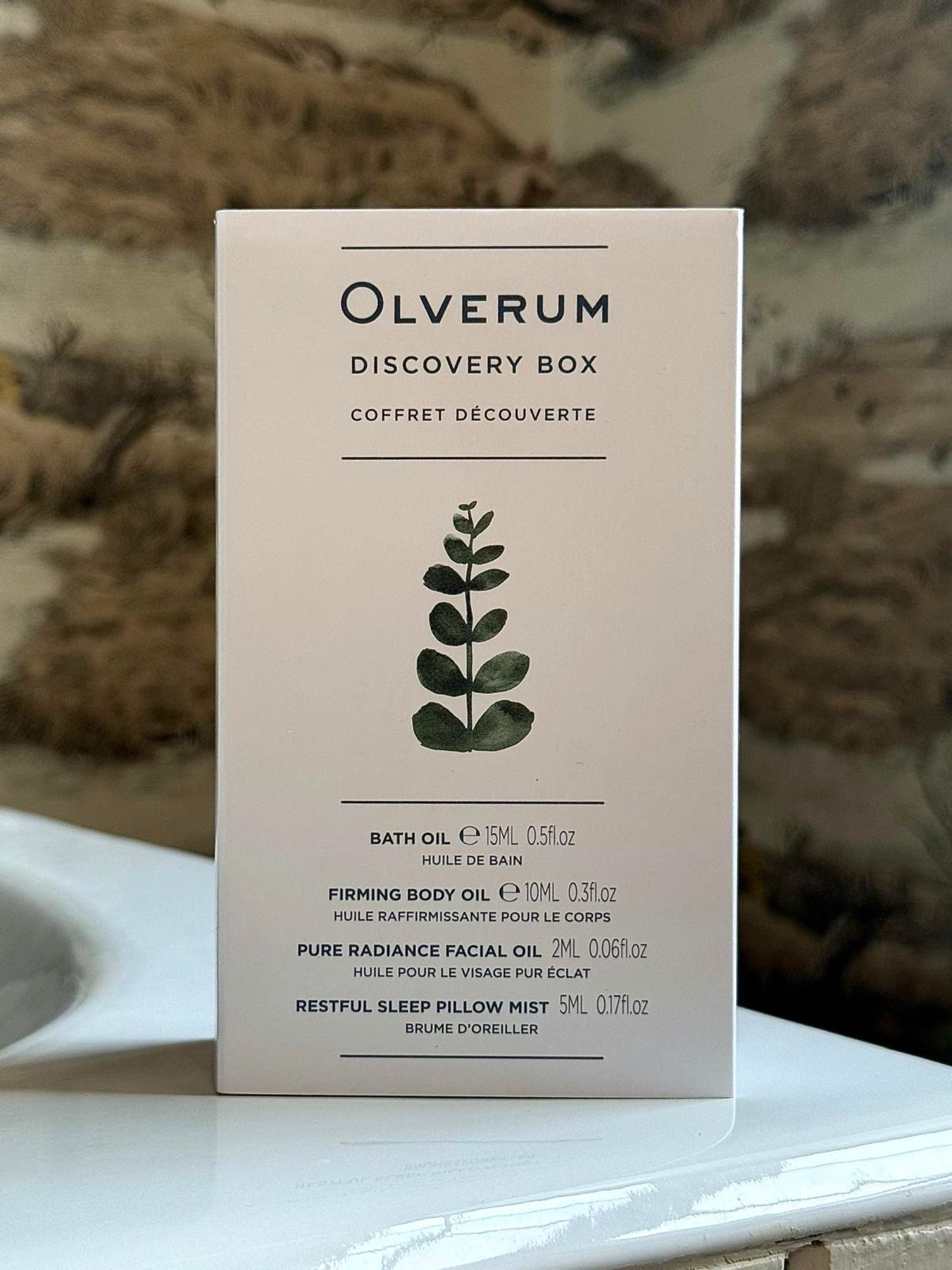 The OLVERUM Discovery Box stands upright with a minimalist design and green botanical illustration, listing included products: bath oil, firming body oil, facial oil, and sleep pillow mist.