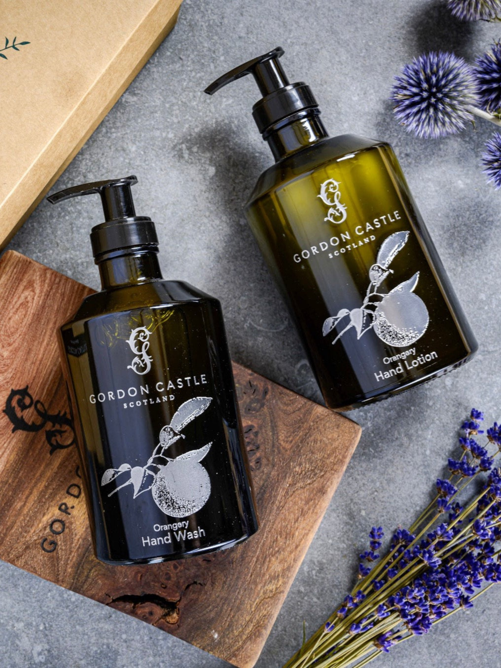 Two refillable Gordon Castle Orangery Hand Lotion glass bottles sit on a wooden board with lavender and thistle, highlighting sustainable luxury next to a cardboard box.