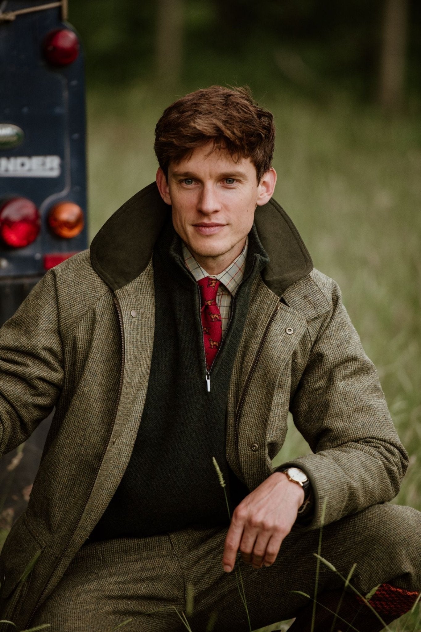A man sits outdoors on grass beside a blue Land Rover, wearing a green tweed jacket, plaid shirt, and Campbells of Beauly Setter Dog Silk Tie. He has short brown hair and looks at the camera with a slight smile.