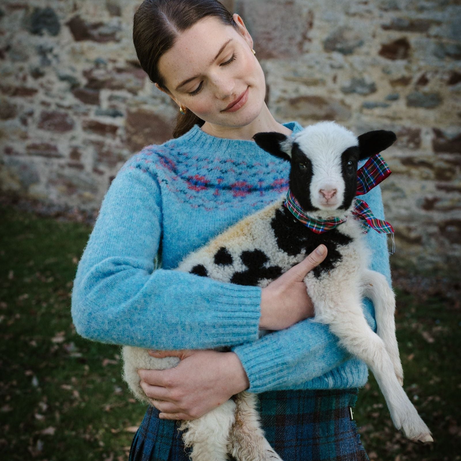A woman in the Shaggy Cropped Fairisle Crew by Campbell's of Beauly stands outside by a stone wall, gently holding a black-and-white lamb with a plaid ribbon, gazing down at it with an affectionate smile.