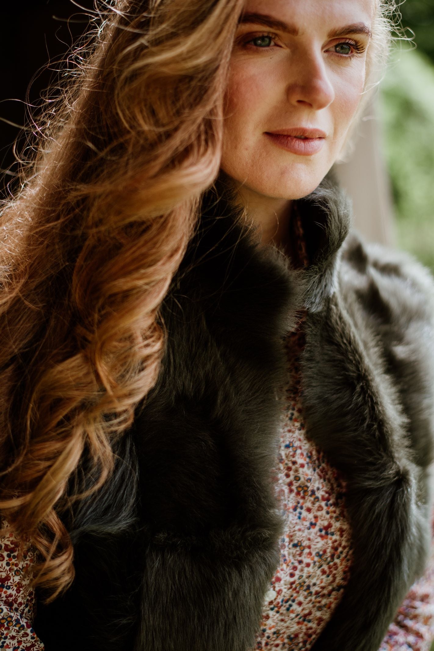 A woman with long, wavy auburn hair wears the Campbells of Beauly Sheepskin Reversible Gilet over a floral top, standing indoors as soft natural light gently highlights her face.