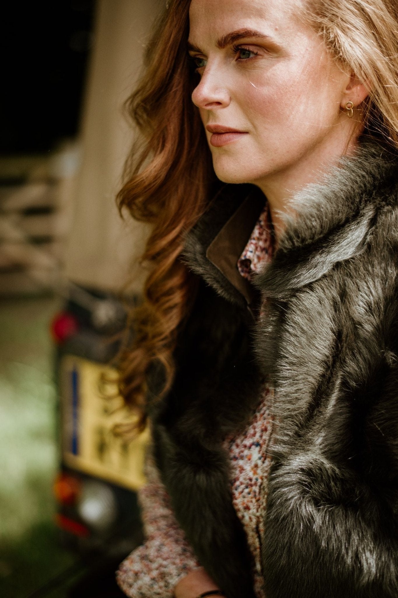 A woman with long, wavy auburn hair wears the Campbells of Beauly Sheepskin Reversible Gilet over a patterned shirt, gazing thoughtfully outdoors with a blurred vehicle and greenery in the background.