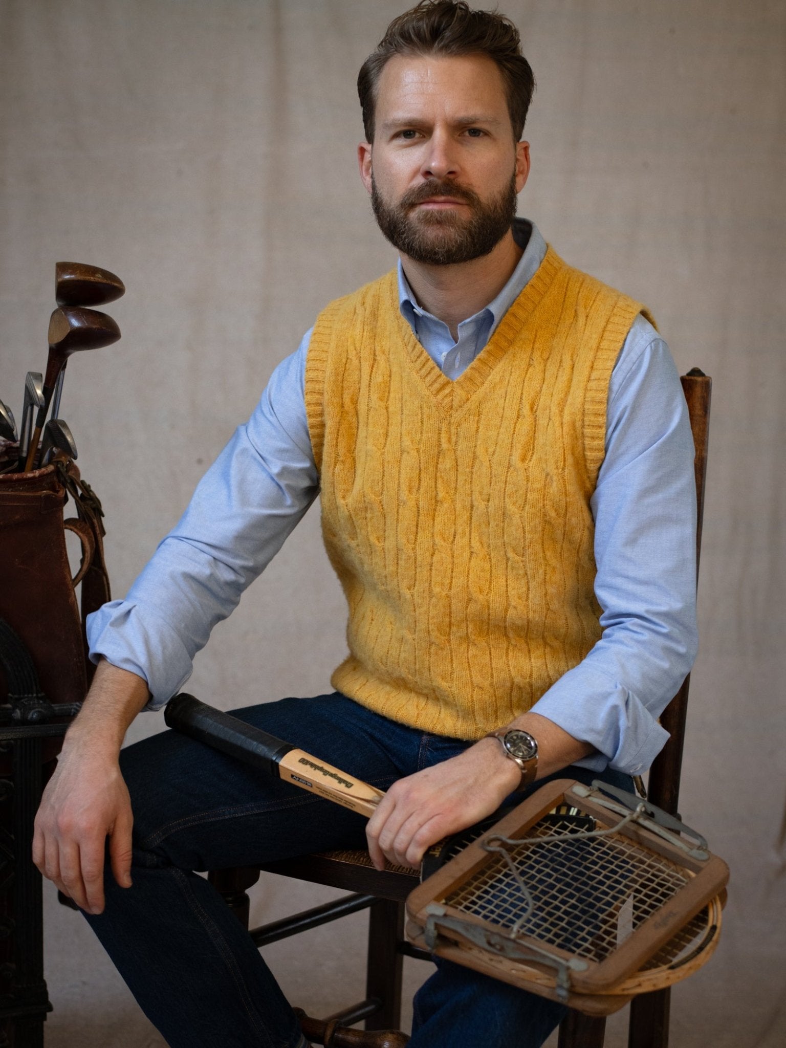 A bearded man sits on a chair in a blue shirt and Campbell's of Beauly Shetland Cable Slipover, holding a broken tennis racket. A golf bag with clubs stands beside him.