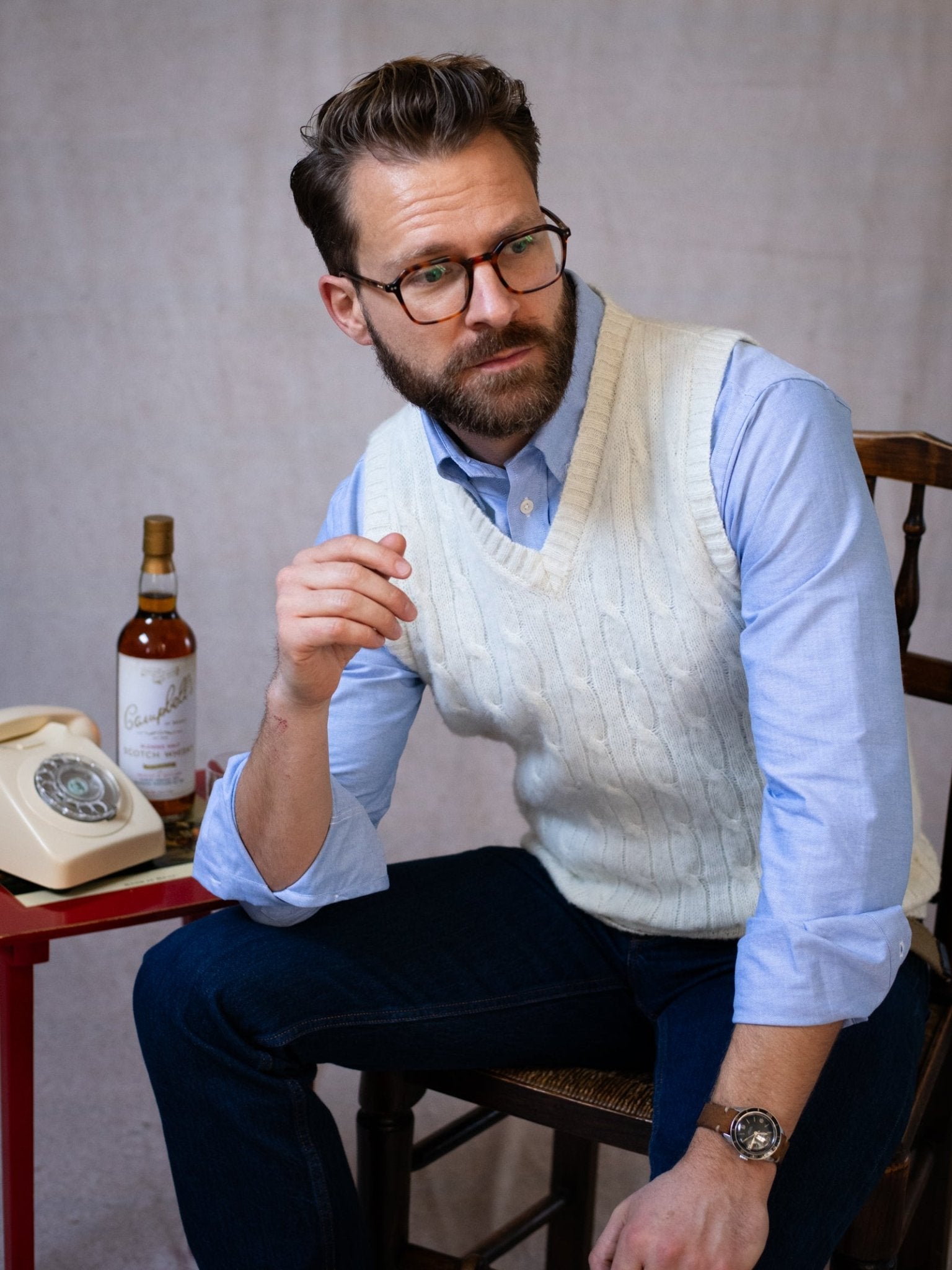 A man with glasses, a beard, and styled hair sits on a chair wearing the Campbell's of Beauly Shetland Cable Slipover over a light blue shirt. A rotary phone and bottle rest on a small red table beside him.