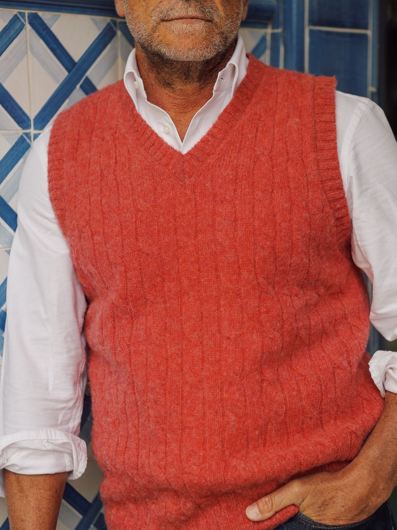A man wearing a white dress shirt and a red Campbell’s of Beauly Shetland Cable Slipover stands in front of a blue and white tiled wall, with one hand in his pocket.
