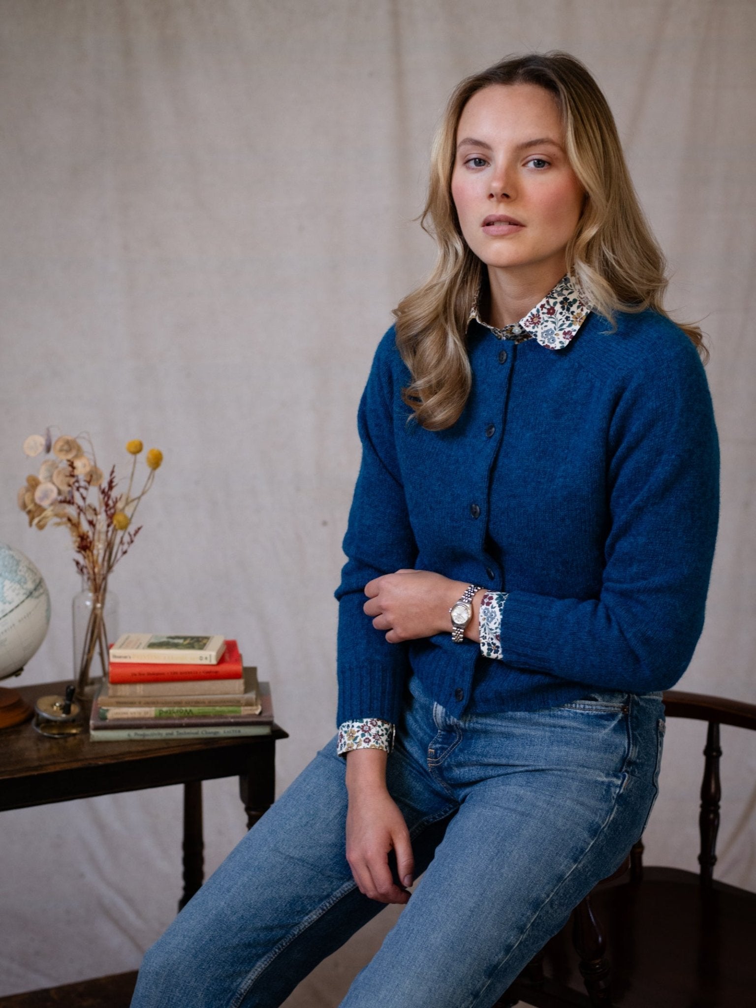 A woman with wavy blonde hair, wearing the Shetland Cropped Cardigan in Azure by Campbell's of Beauly and jeans, sits on a wooden chair beside a small table with a globe, dried flowers, and stacked books against a neutral background.