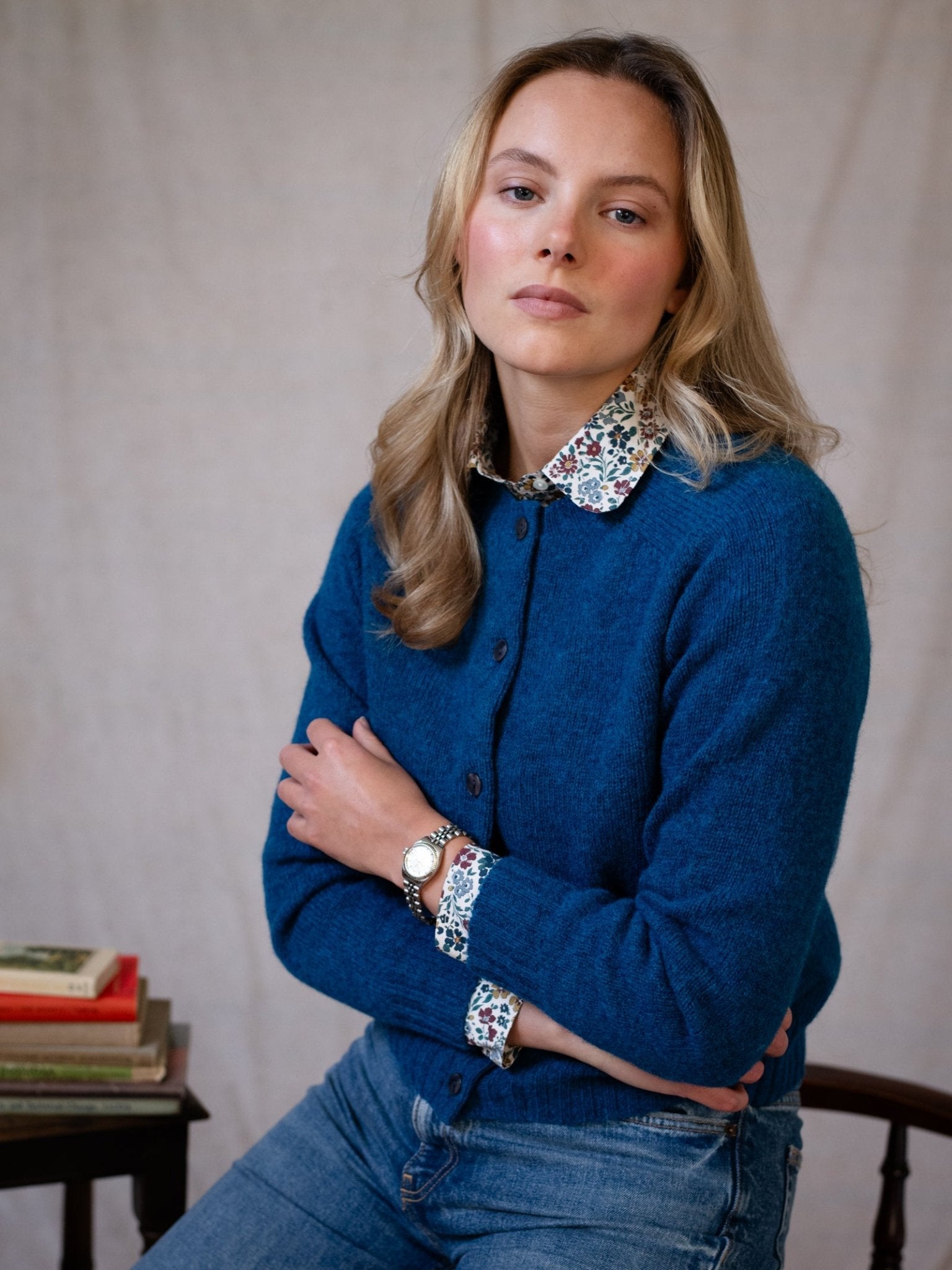 A woman with long blonde hair sits on a chair, calmly looking at the camera. She wears the Shetland Cropped Cardigan by Campbell's of Beauly over a floral shirt and blue jeans, with her arms folded and a stack of books beside her.