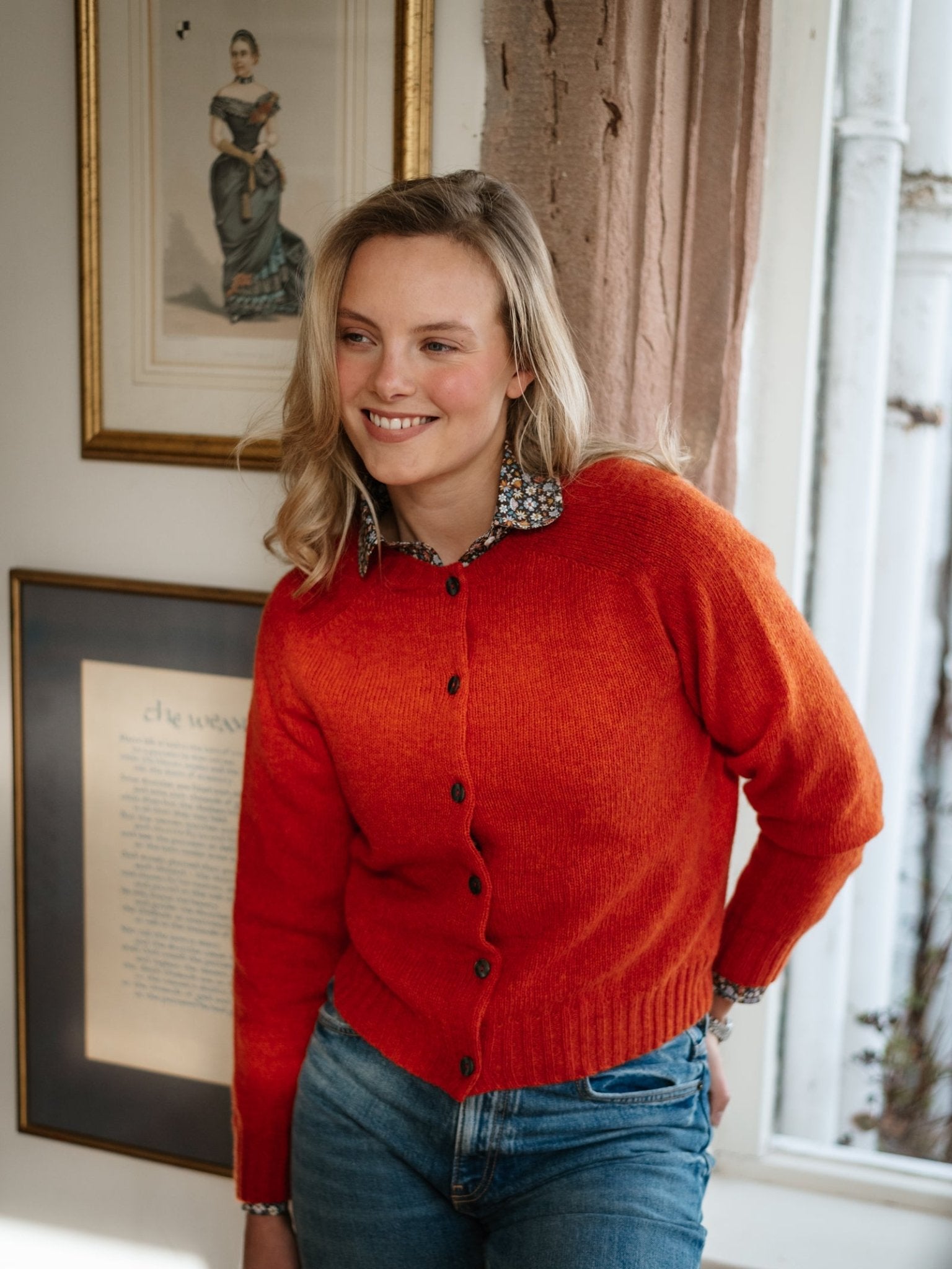 A smiling woman wears a bright orange Shetland Cropped Cardigan by Campbell's of Beauly, made from Pure New Wool, standing indoors near framed artwork and a window with one hand on her hip.