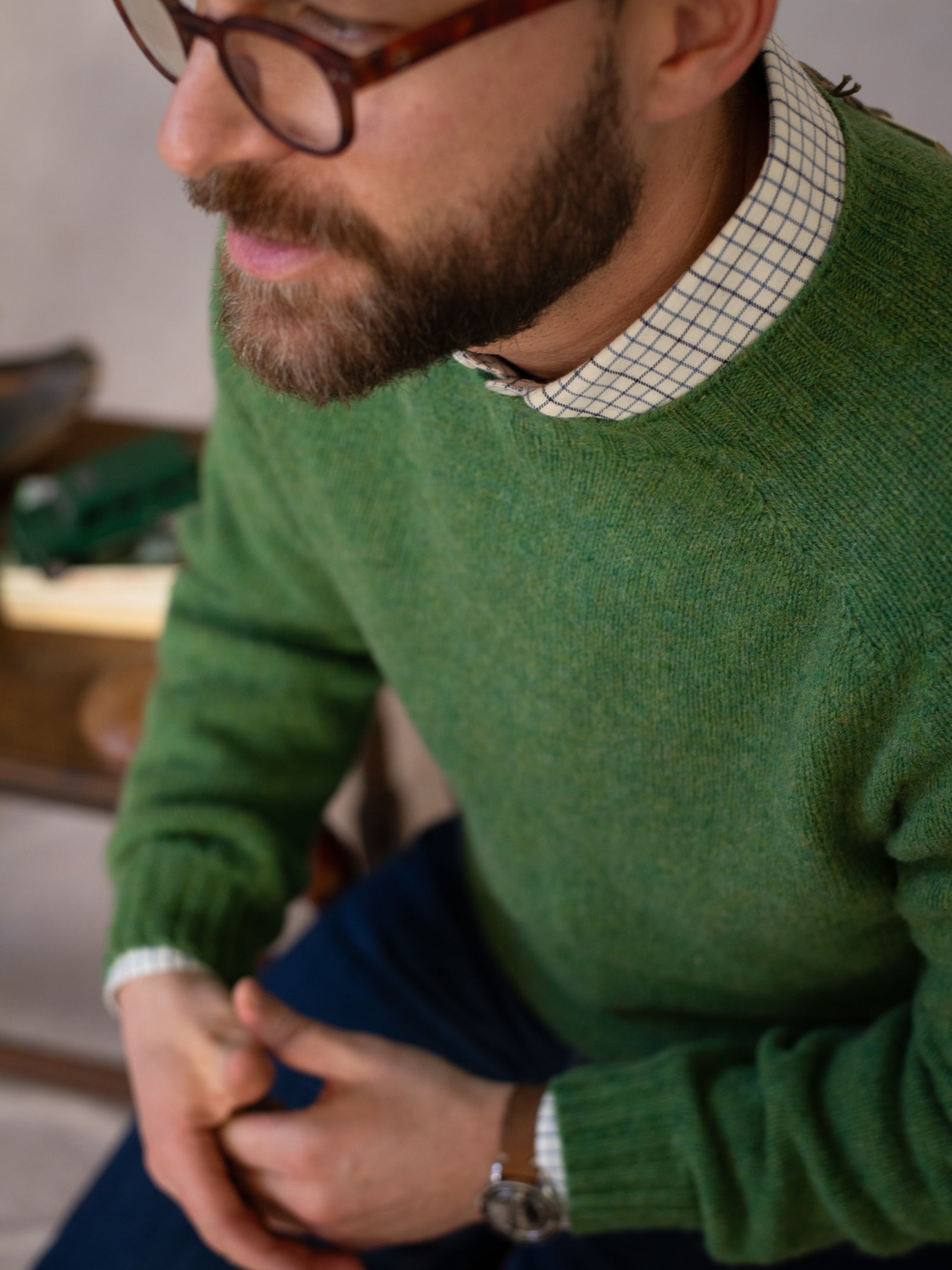 A bearded man in glasses wears a green Campbell's of Beauly Shetland Jumper over a checkered shirt and blue jeans, sitting with his hands together, visible from the chest down.