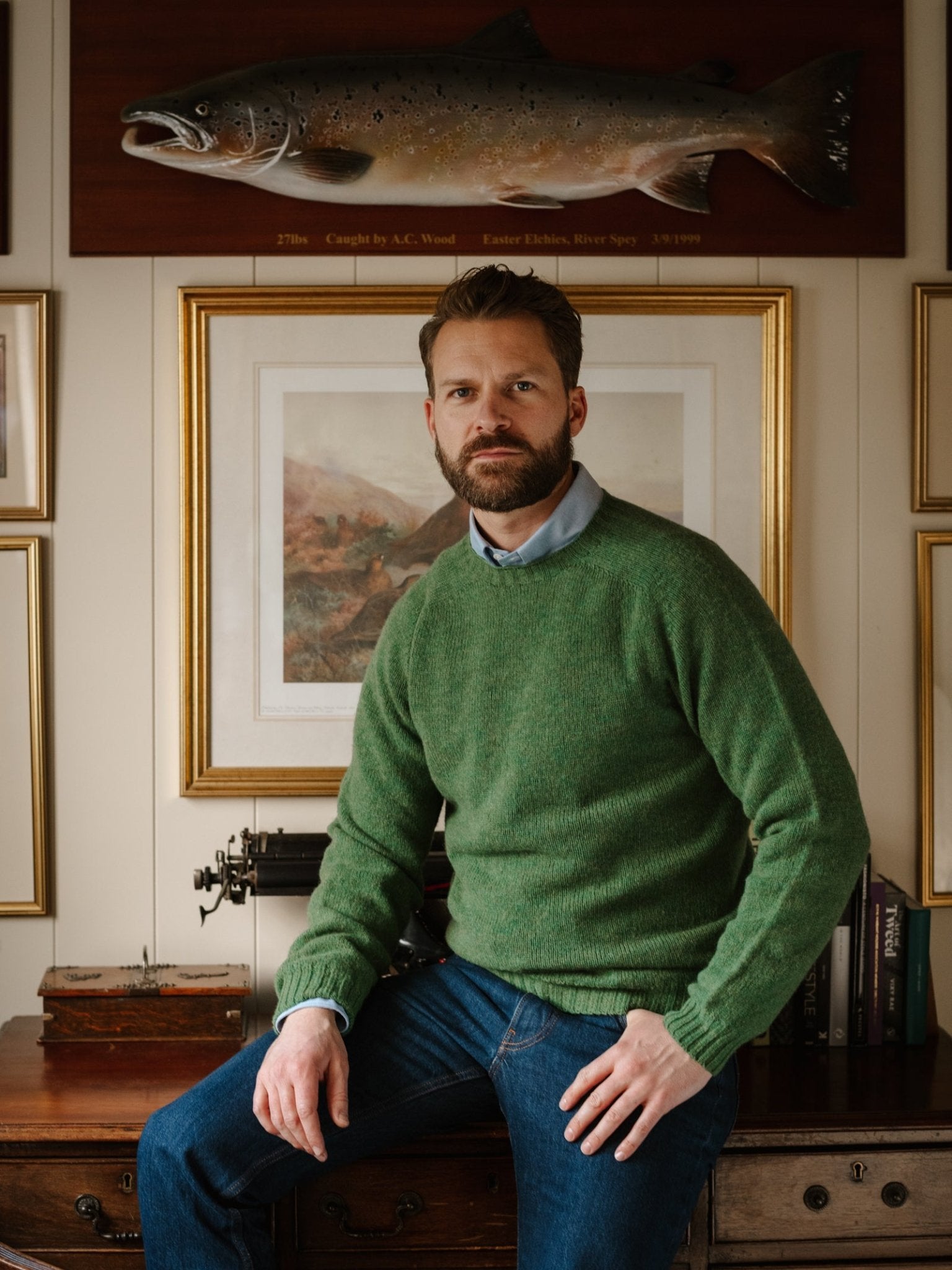 A bearded man, dressed in a green Campbell's of Beauly Shetland Jumper and blue jeans, sits on a wooden desk in a room with framed artwork and a mounted fish on the wall behind him.