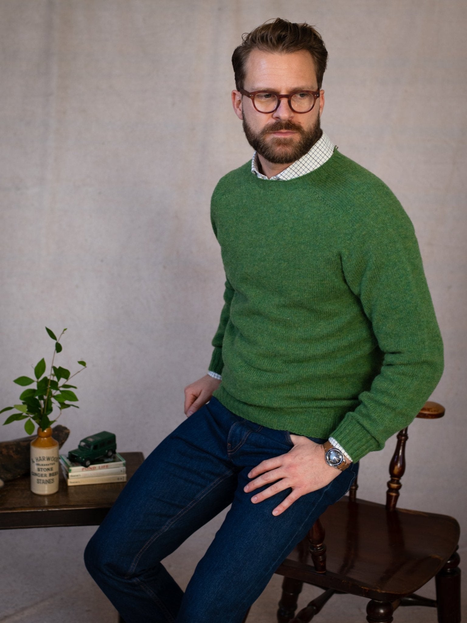 A bearded man with glasses, wearing a green Campbell's of Beauly Shetland Jumper and blue jeans, sits on a wooden chair next to a small table with books, a potted plant, and a model car. The background is plain and neutral.
