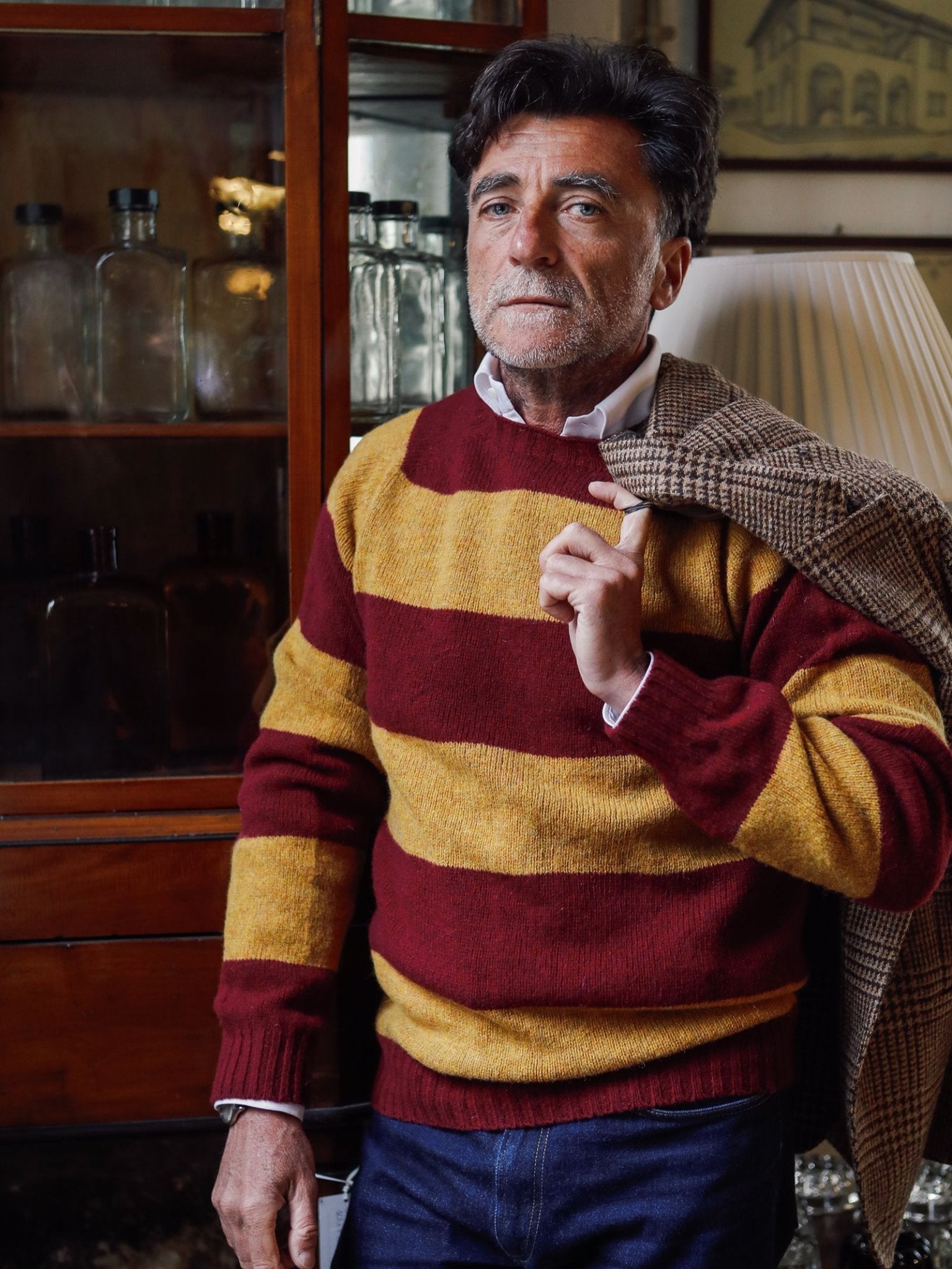A man with gray hair and a beard wears the Campbell’s of Beauly Shetland Stripe Crew Jumper in red and yellow, holding a plaid jacket over his shoulder. He stands indoors near glass bottles and a lamp, highlighting the timeless appeal of this heritage knit crafted from pure Shetland lambswool.