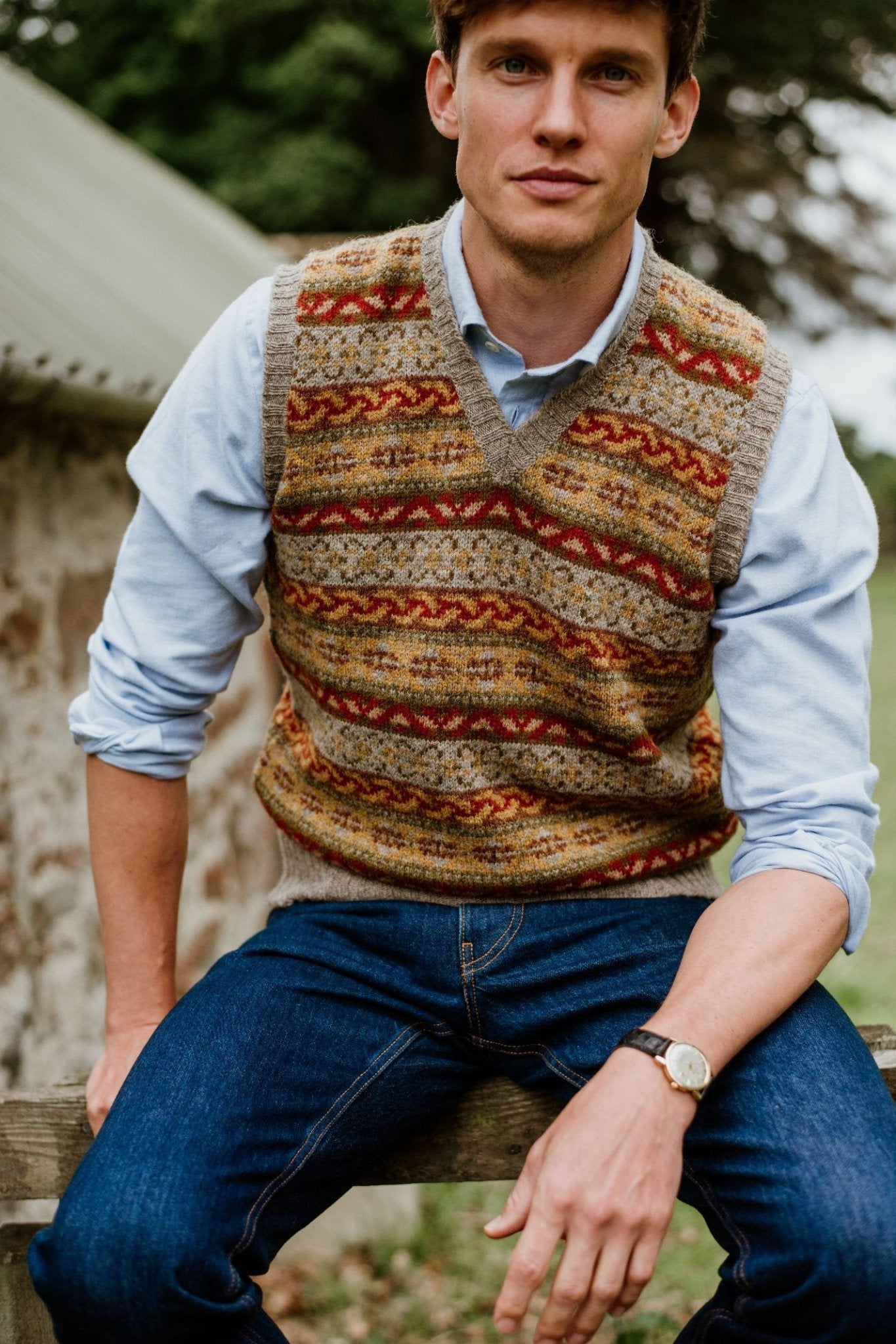 A man sits outdoors on a wooden fence, wearing the Campbells of Beauly Fairisle Slipover over a light blue shirt and blue jeans, with a watch on his left wrist. A stone building and greenery form the background.