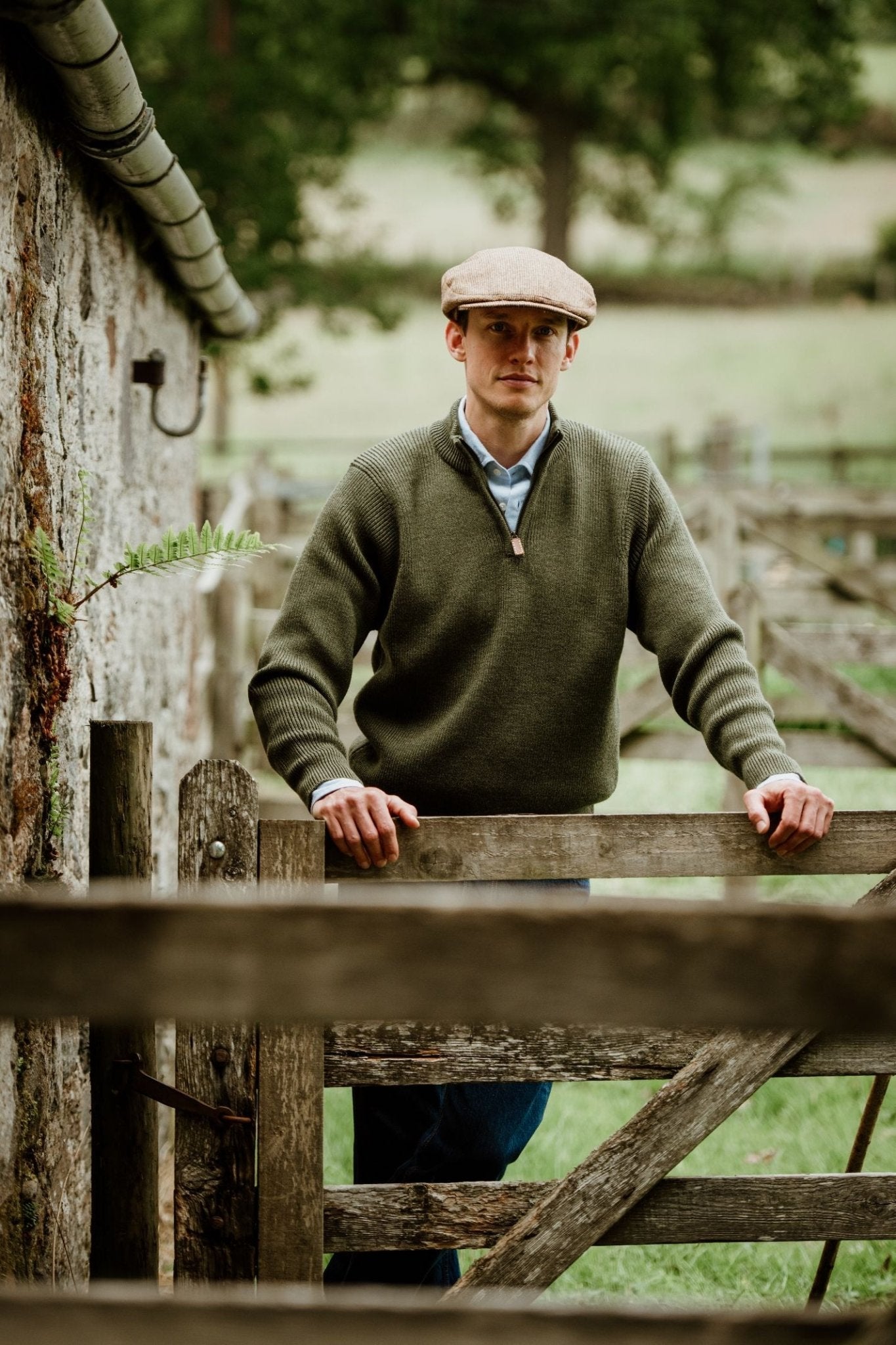 A man wears a Campbells of Beauly Shooter Quarter Zip Jumper with Patches, blue shirt, and jeans, standing outdoors by a wooden gate and stone wall, with grassy fields and trees in the background.