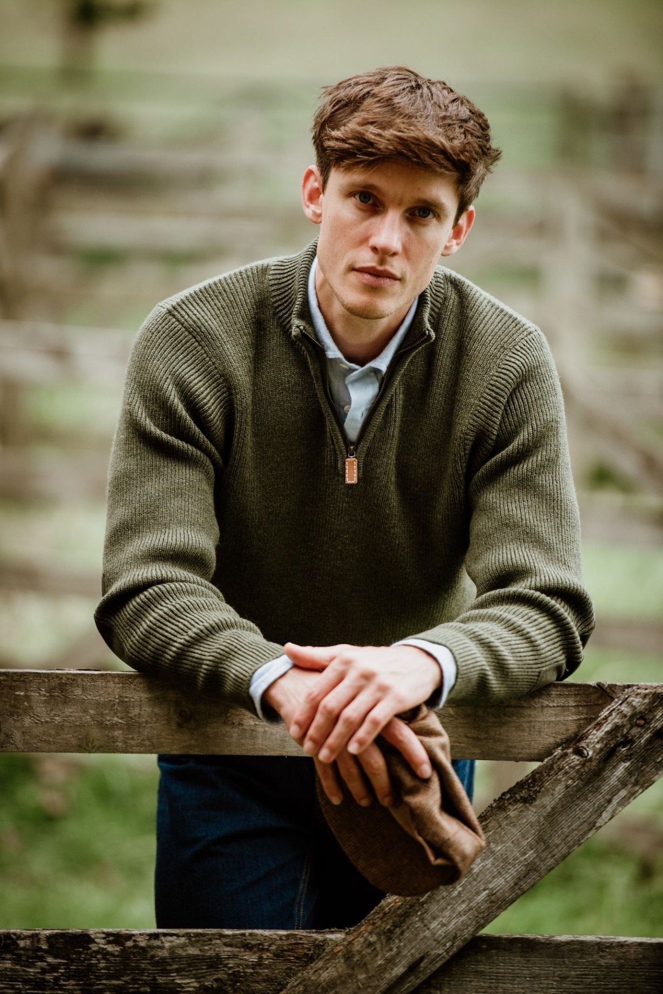 A young man with short brown hair leans on a wooden fence outdoors, wearing Campbells of Beauly Shooter Quarter Zip Jumper with Patches over a collared shirt and holding a brown flat cap. The grassy background is blurred.