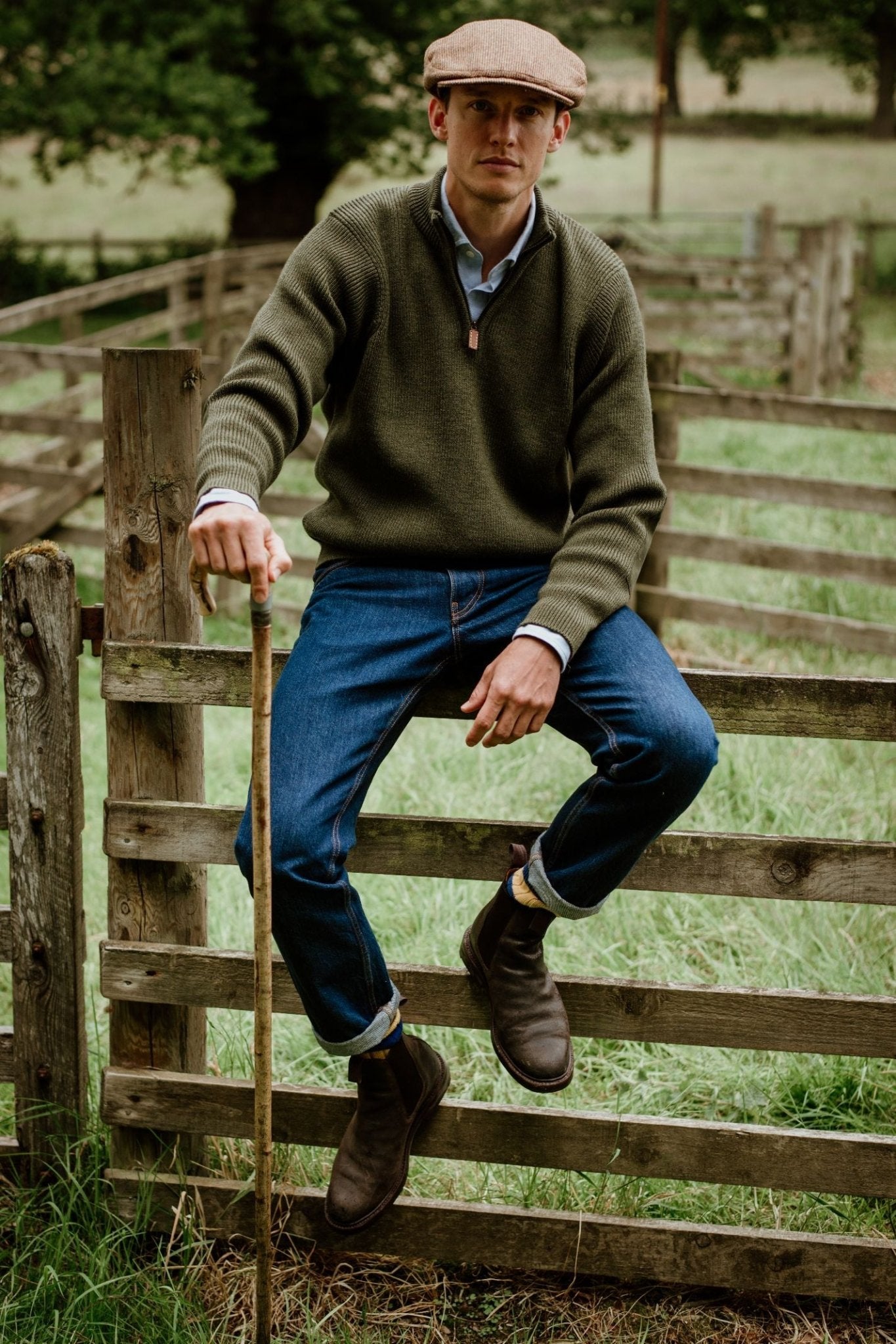 A man in a flat cap, Campbells of Beauly Shooter Quarter Zip Jumper with Patches, blue jeans, and brown boots sits on a wooden fence in a grassy outdoor setting, holding a walking stick and looking at the camera.
