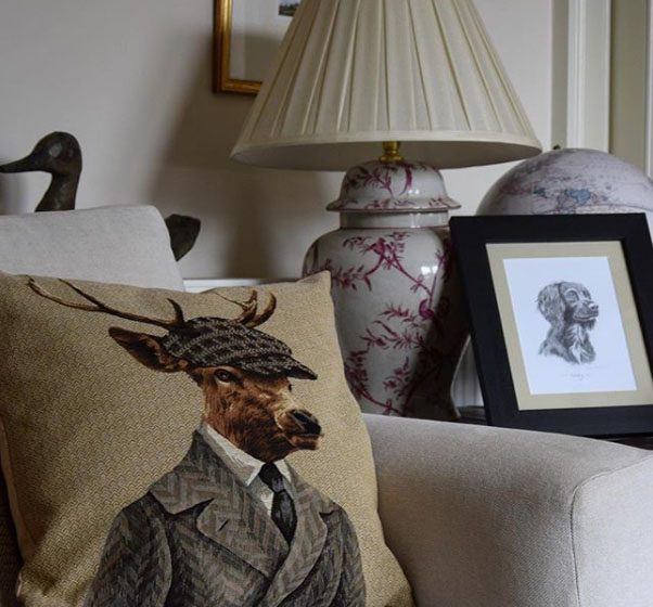 A beige couch showcases the Dapper Stag Cushion Cover by Campbells of Beauly. Nearby, a table displays a floral-based lamp, globe, framed dog sketch, and wooden bird sculpture for cozy, eclectic decor.