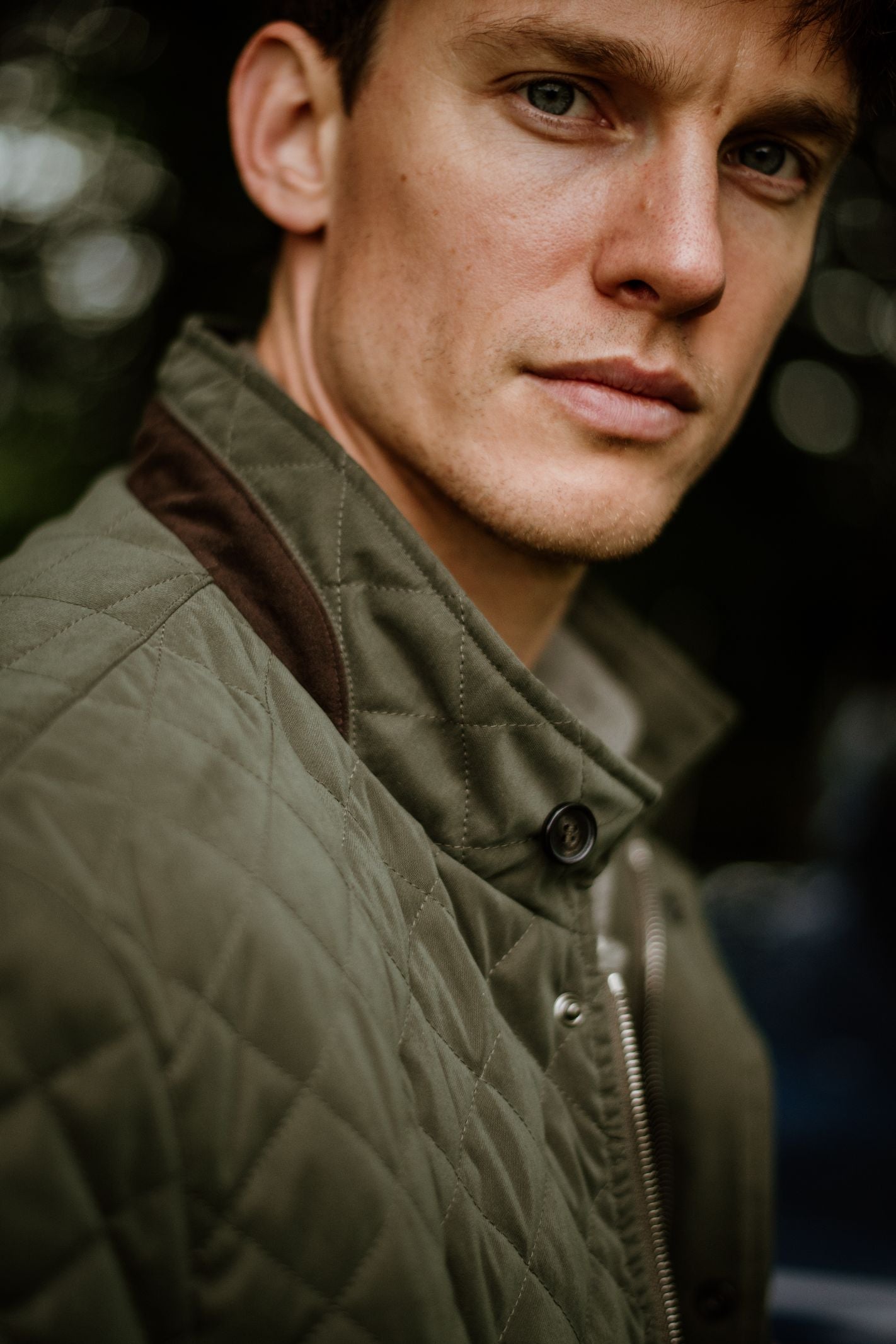 A young man with short brown hair and blue eyes looks at the camera, wearing the Tomich Jacket by Campbells of Beauly—a green quilted coat with a brown collar. The blurred background features hints of greenery.