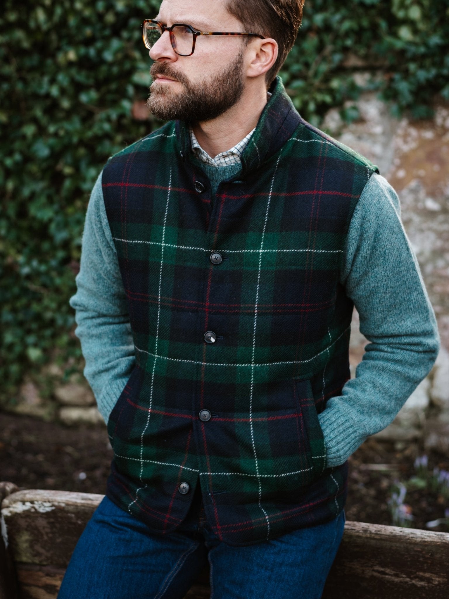 A bearded man in glasses and jeans sits outdoors before a leafy wall, wearing a blue-green sweater and the Campbell's of Beauly Auld Stock Quilted Tweed Buttoned Gilet, looking to the side with his hands on his thighs.
