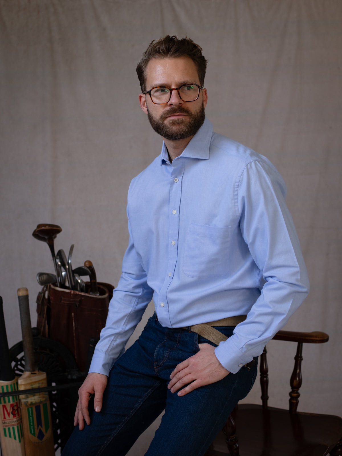 A man with glasses and a beard, dressed in the Campbell’s of Beauly Twill Shirt in light blue classic fit and jeans, sits on a wooden chair. Behind him are a vintage golf bag with clubs and a cricket bat, set against a plain beige cloth backdrop.