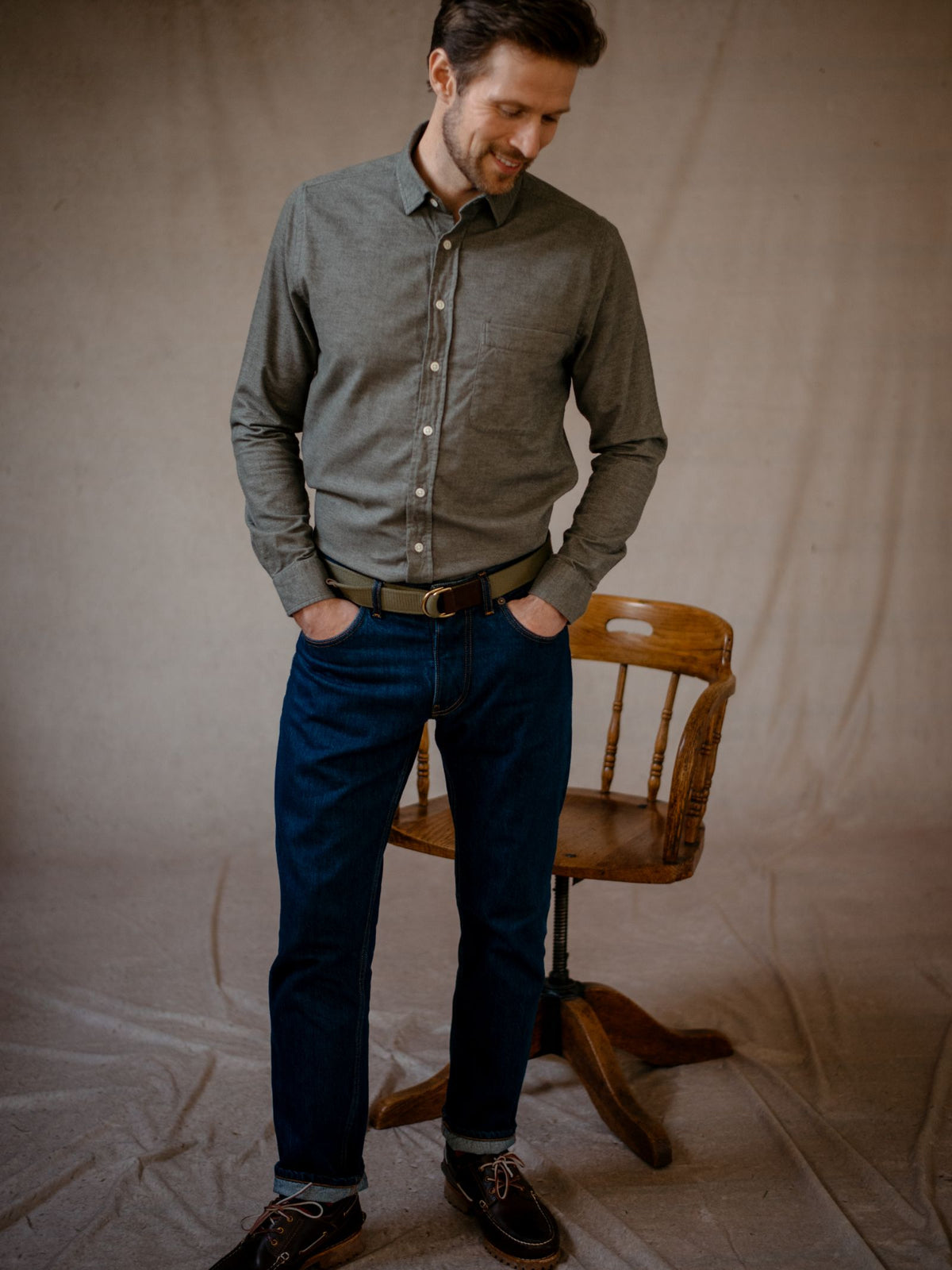 A man wearing the Campbell’s of Beauly Olive Twill Shirt and blue jeans stands with his hands in his pockets, looking down and smiling. A wooden chair is positioned behind him on a fabric-covered floor.