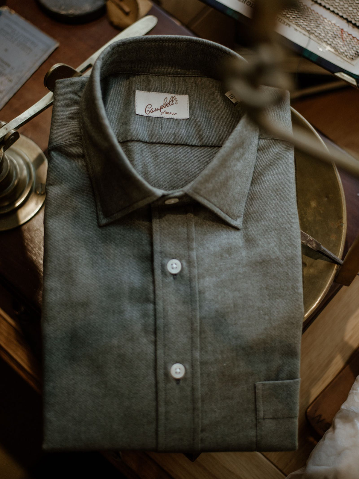 A neatly folded gray Twill Shirt from Campbell’s of Beauly, featuring white buttons, rests on a wooden surface beside a brass lamp.