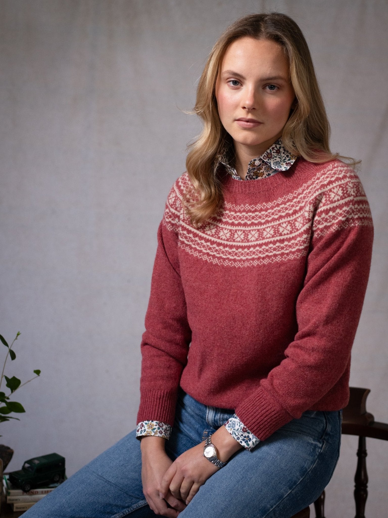 A young woman with wavy blonde hair wears Campbell's of Beauly's Two-Colour Fairisle Yoke Jumper over a floral shirt and blue jeans, seated on a chair and gazing slightly to the side against a neutral background.