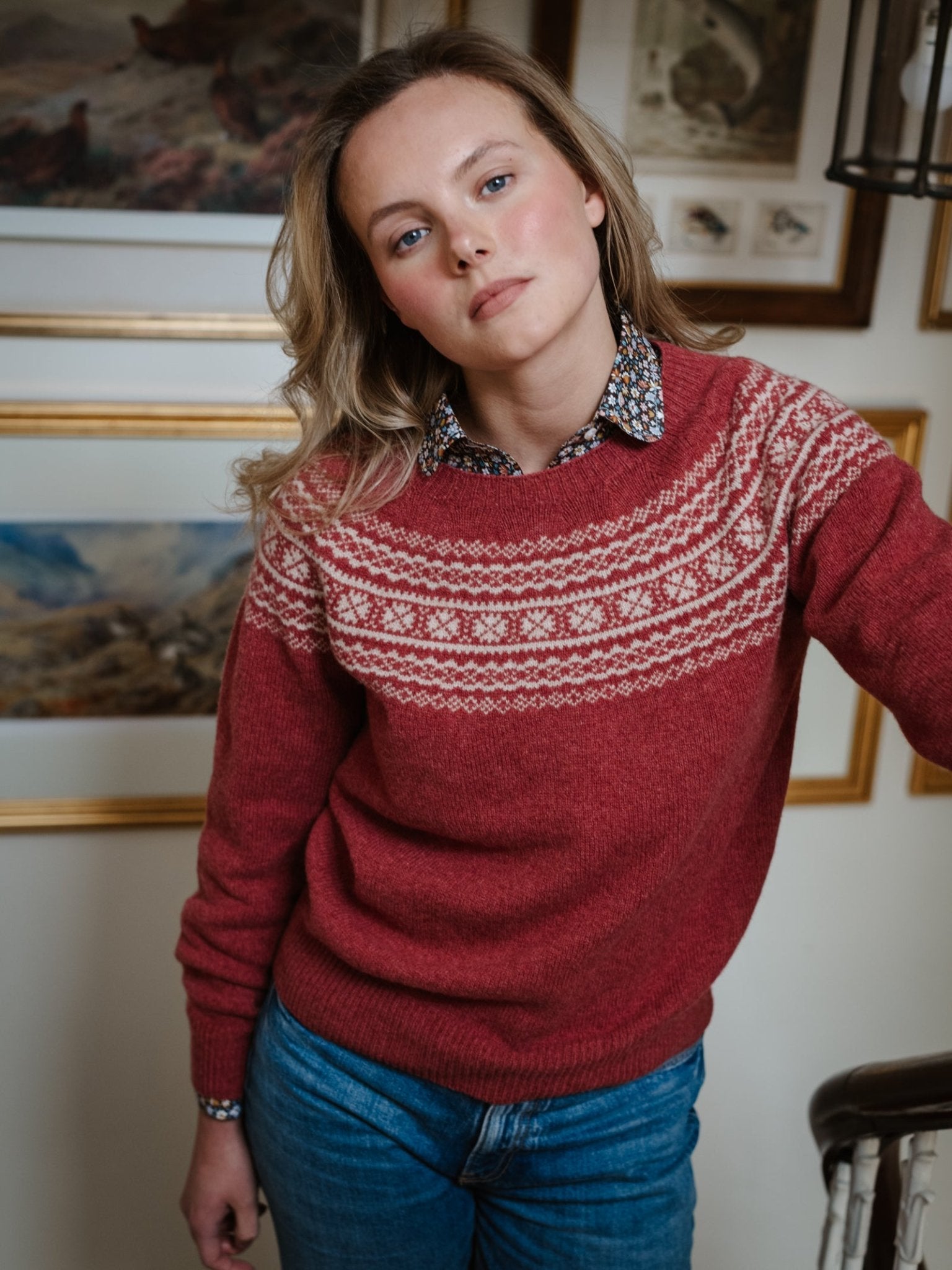 A blonde woman wears a Campbell's of Beauly Two-Colour Fairisle Yoke Jumper in Rosewood and jeans, standing indoors before framed pictures, looking at the camera with a neutral expression.