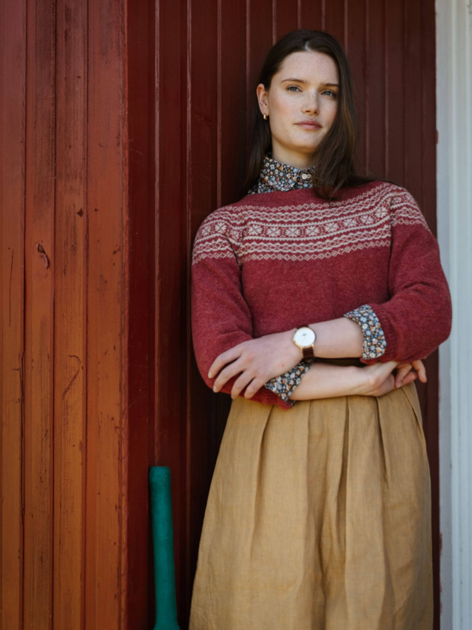 A woman with long brown hair stands against a red wooden wall, wearing Campbell's of Beauly's Two-Colour Fairisle Yoke Jumper over a floral blouse and beige skirt. She crosses her arms and looks at the camera; a green-handled object leans nearby.